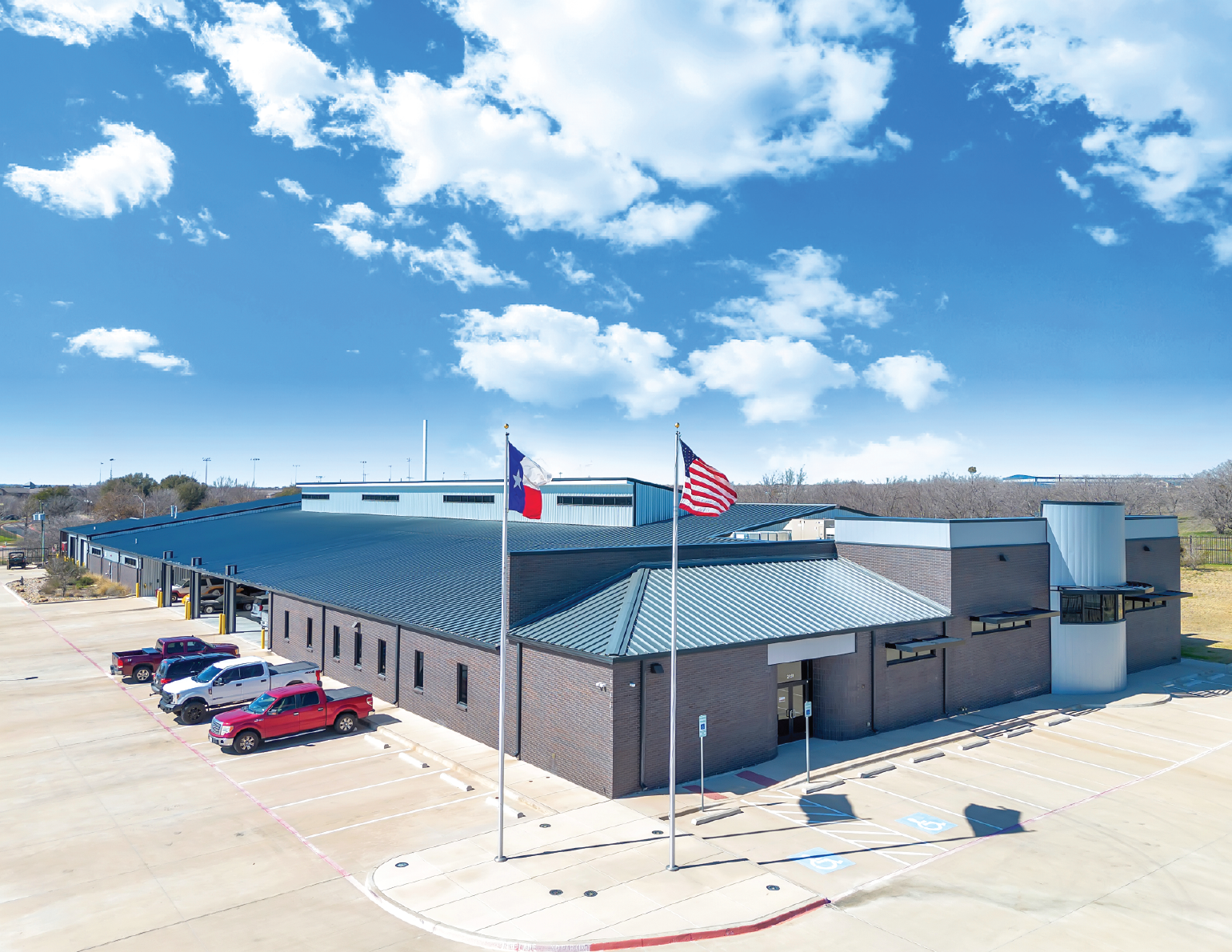 An aerial view of a modern brick building with a metal roof, parking lot, and two flags on flagpoles (Texas and US) against a partly cloudy blue sky.