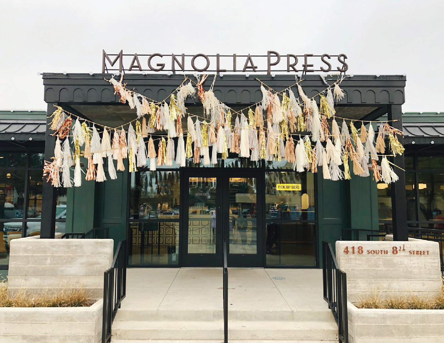 Exterior view of Magnolia Press coffee shop with decorative tassel garlands hanging above the entrance, black frames around glass doors, and an address sign on the right side reading '418 South 8th Street'.