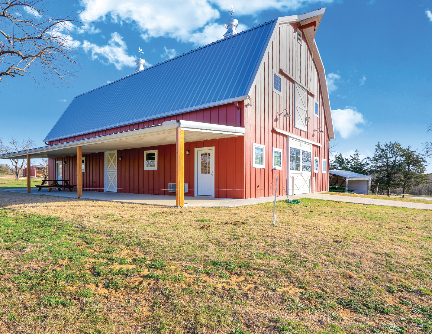 Red barn with white doors, windows, and trim, metal roof, porch with picnic table, trees, and cloudy sky in the background.