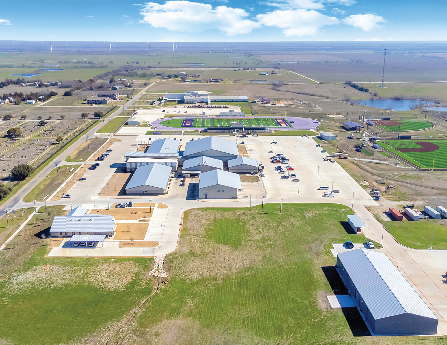 Aerial view of a school or community complex with sports fields, parking lots, and surrounding open land, under a partly cloudy sky.