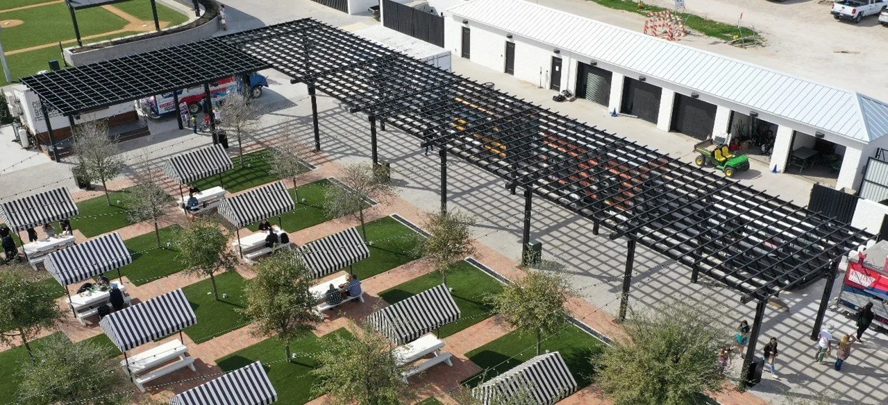 An outdoor area with benches and small trees, partly covered by a black pergola structure, adjacent to a building with a row of garage doors.