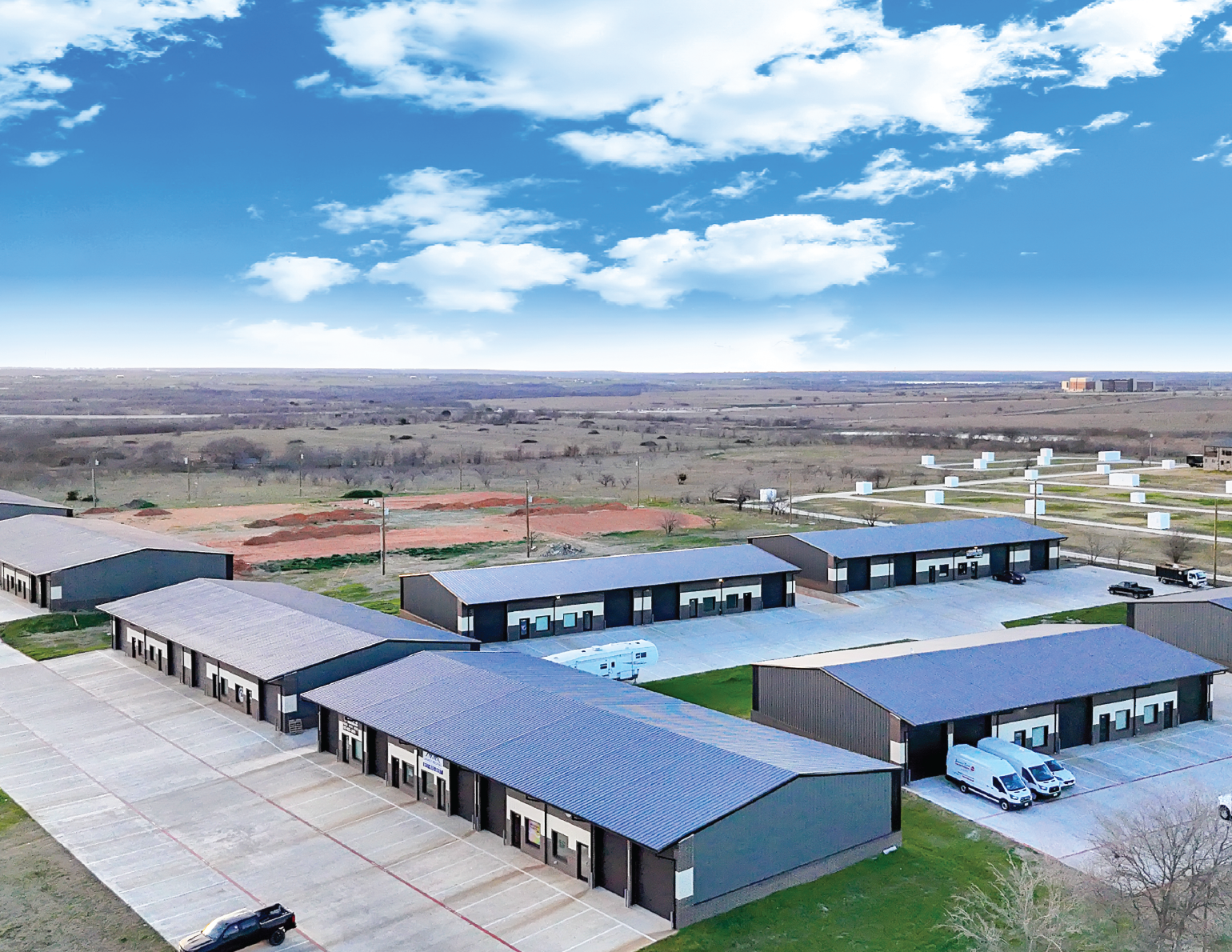 A commercial area with multiple single-story buildings, parking lots, and trucks, set against a rural landscape with open fields and a blue sky with clouds.