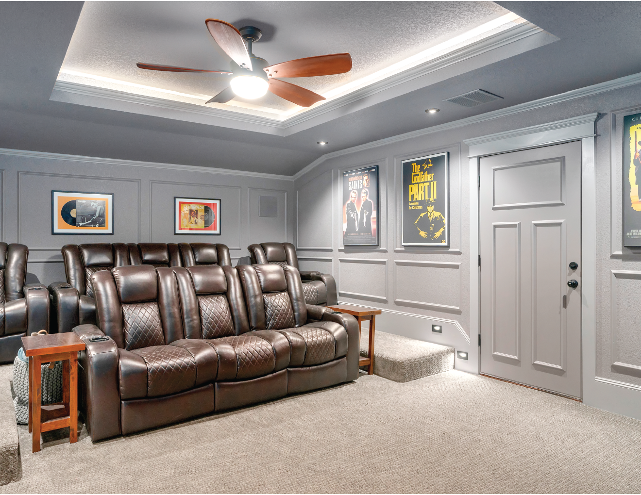 A cozy home theater room with a gray ceiling and walls, featuring framed movie posters, a ceiling fan, and plush brown leather seating arranged in rows on beige carpet.