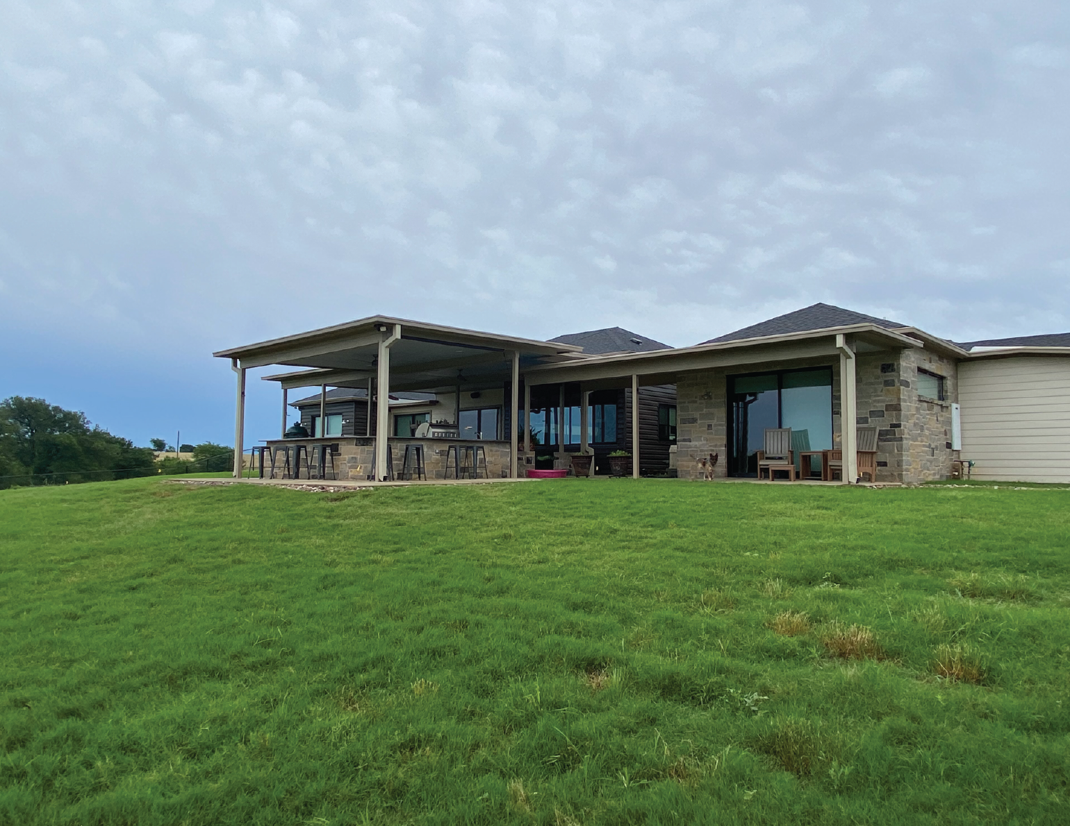 A modern single-story house with a large back porch, stone accents, and sliding glass doors, overlooking a well-maintained grassy yard. The porch has outdoor furniture and bar seating.