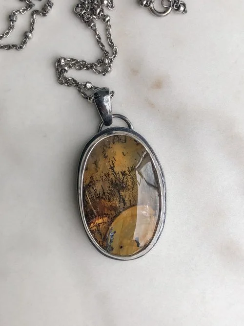 Orangey brown iron oxides fill this oval shaped Quartz stone. The branch-like dendrites rise above what reminds me of a hilltop. The chain features tiny faceted beads. The stone is rests in a hand-dipped, two-toned setting.
