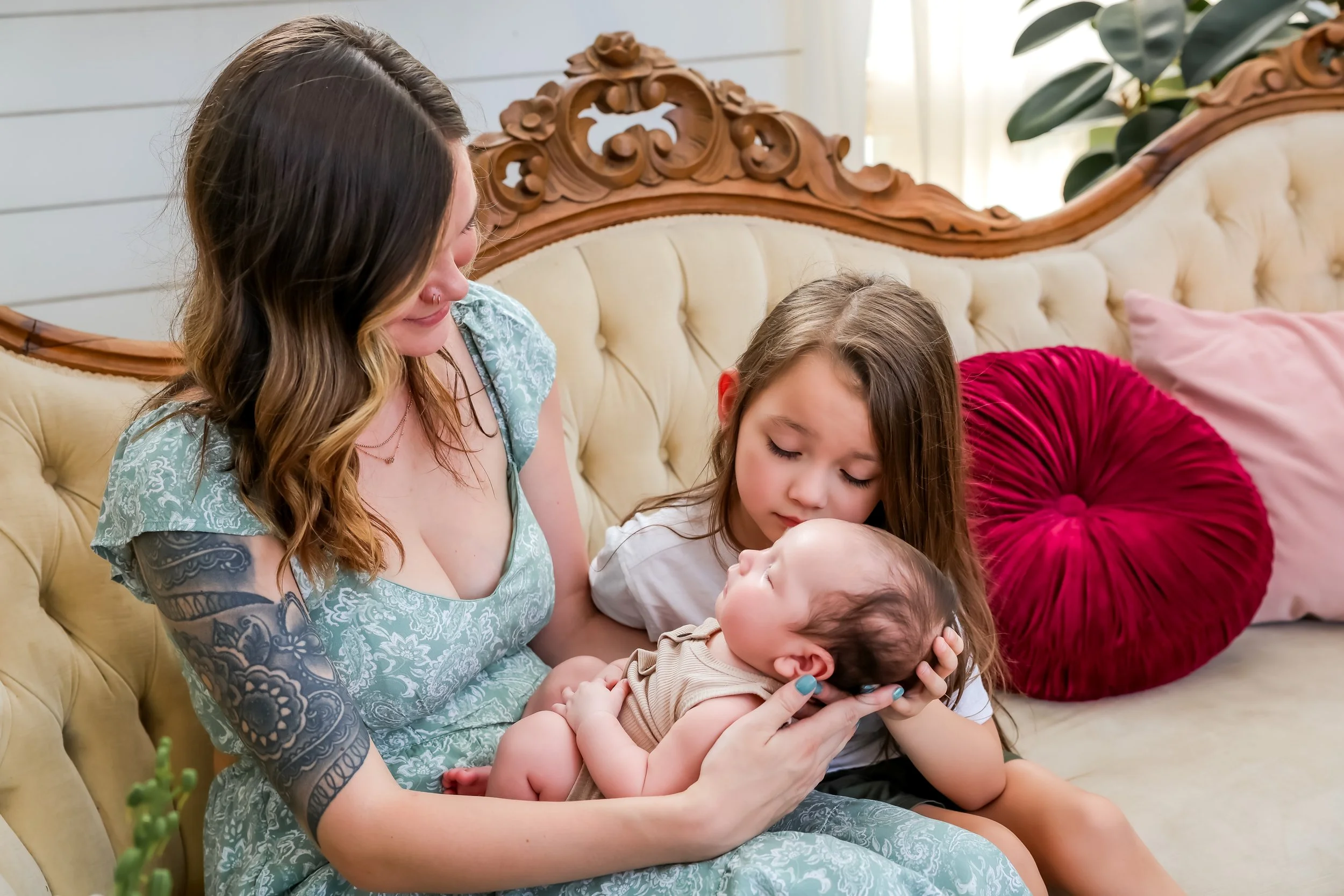 A woman with a floral tattoo on her arm and two young girls sitting on a vintage cream-colored sofa with wooden carvings, holding a baby in her arms, in a warmly lit living room.