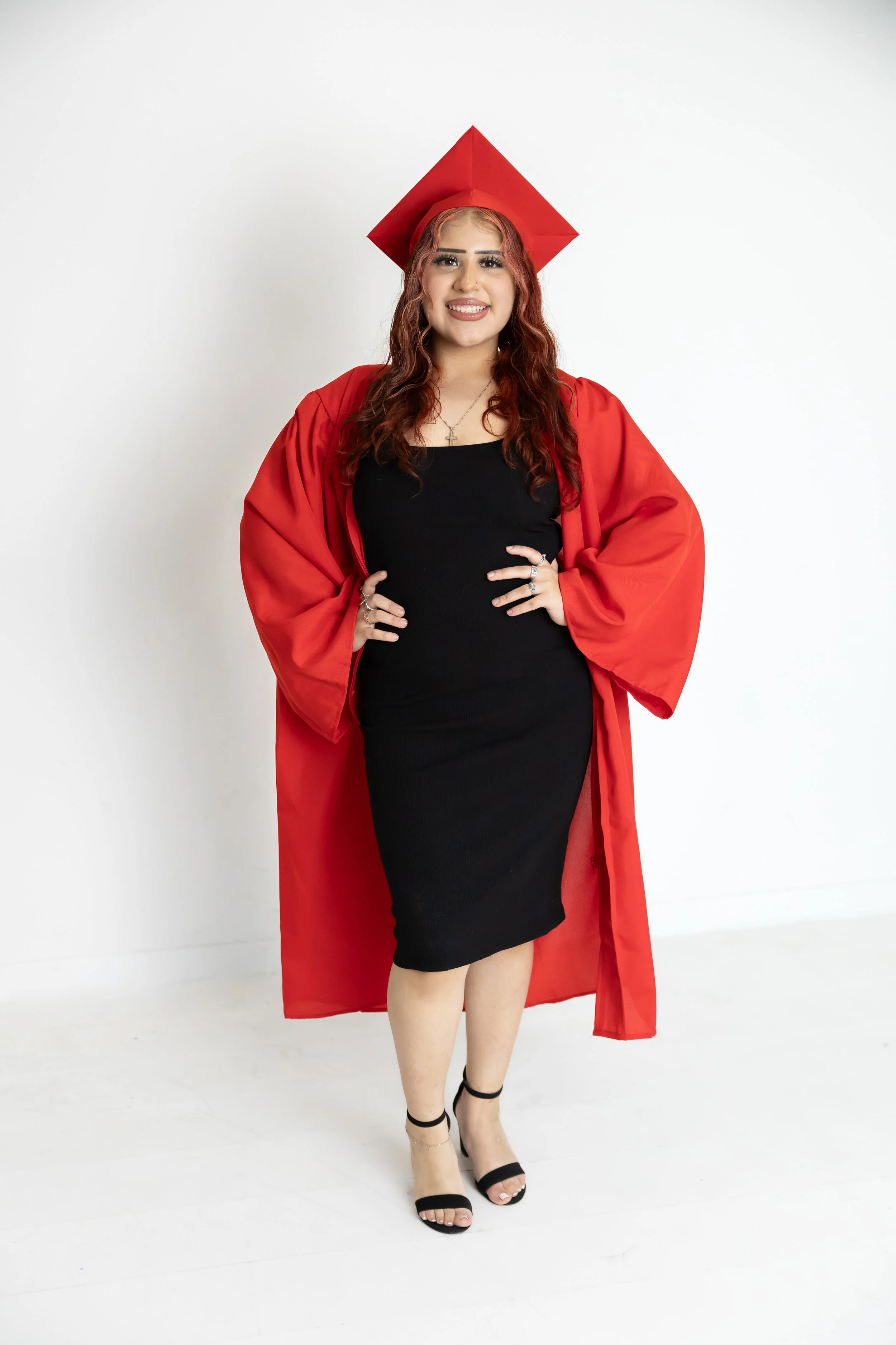 A woman in a red graduation gown and cap, wearing a black dress and black heels, standing against a white background.