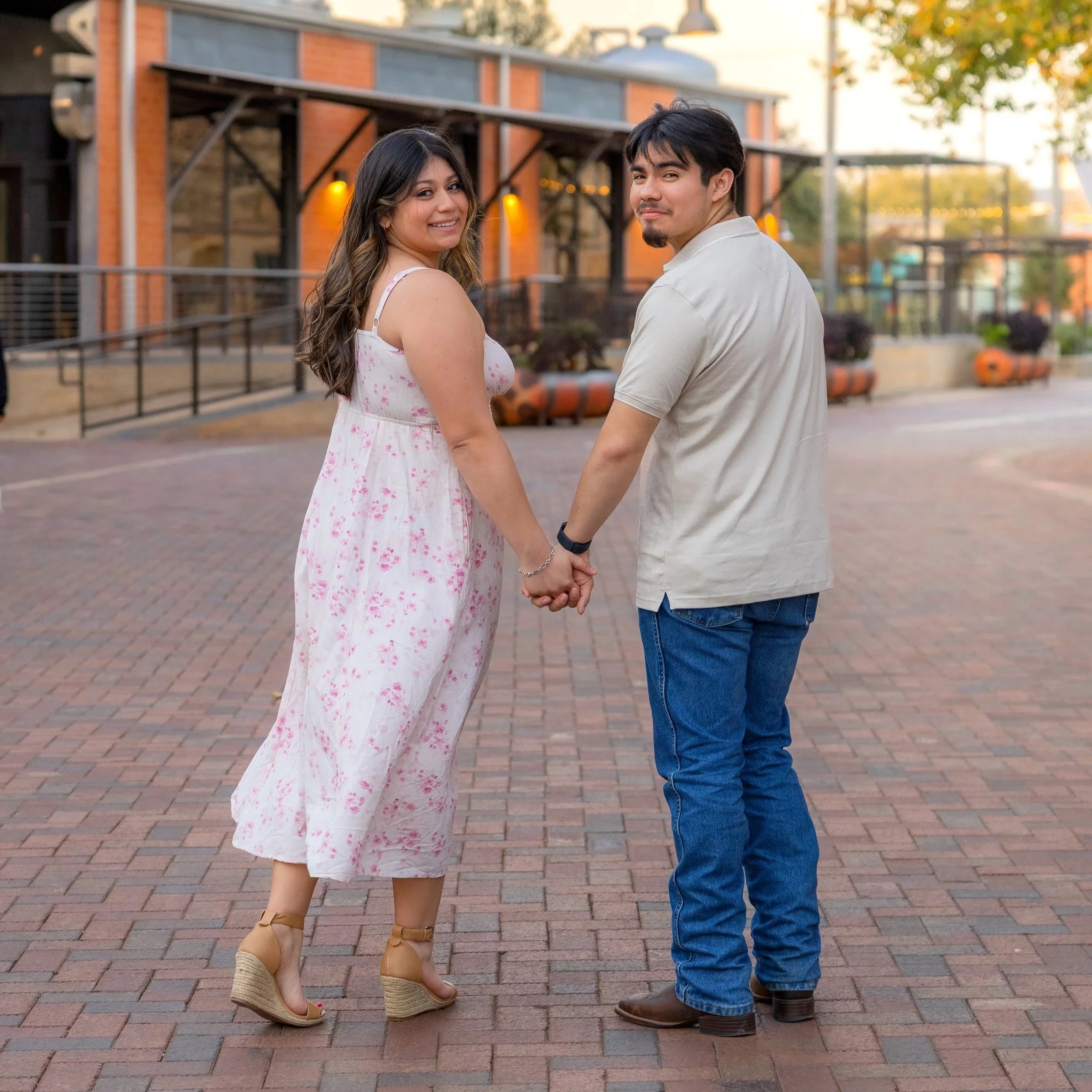A couple holding hands and looking back at the camera outdoors on a brick-paved area, with a building and trees in the background during sunset.