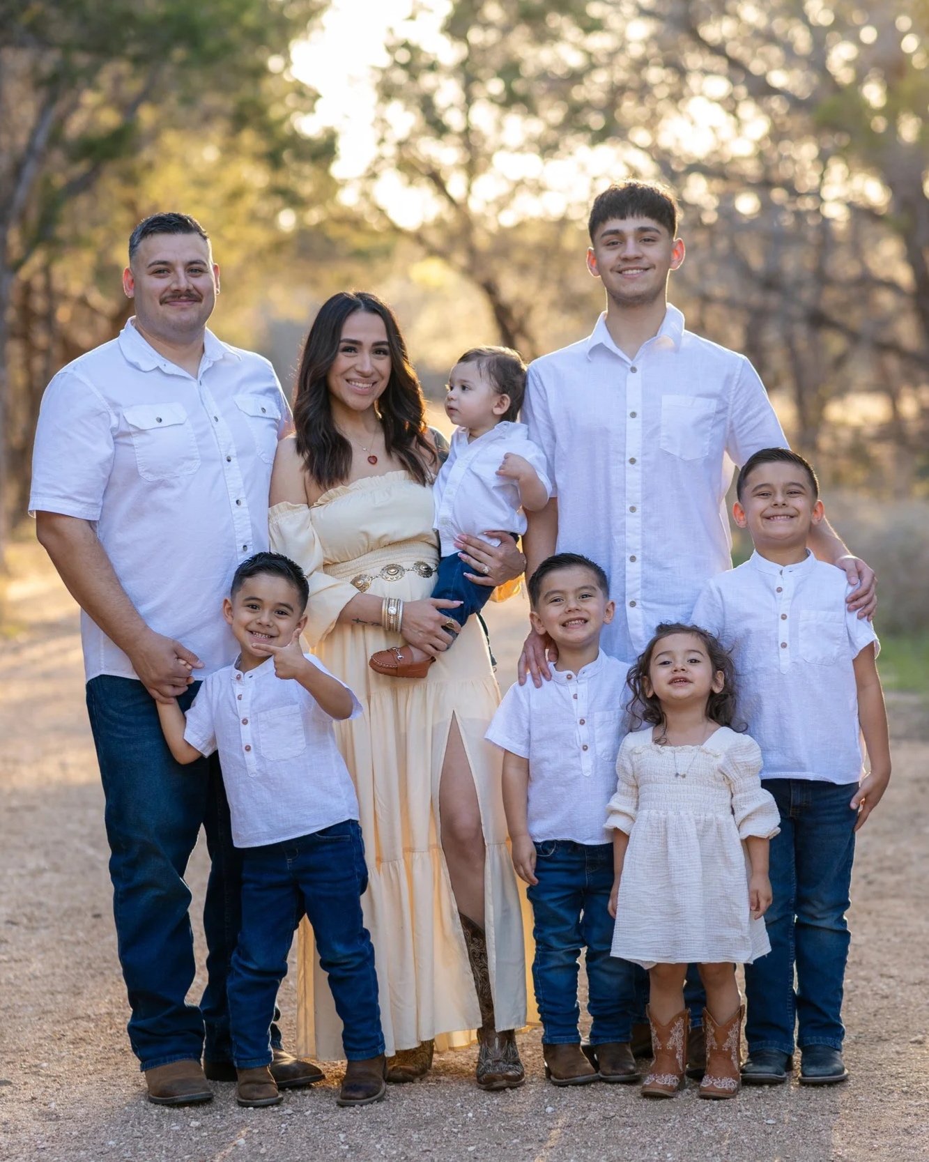 Family of nine outdoors at sunset, posing for a photo, including two adult men, one adult woman, three boys, and two girls, all dressed in casual white and light-colored clothing.