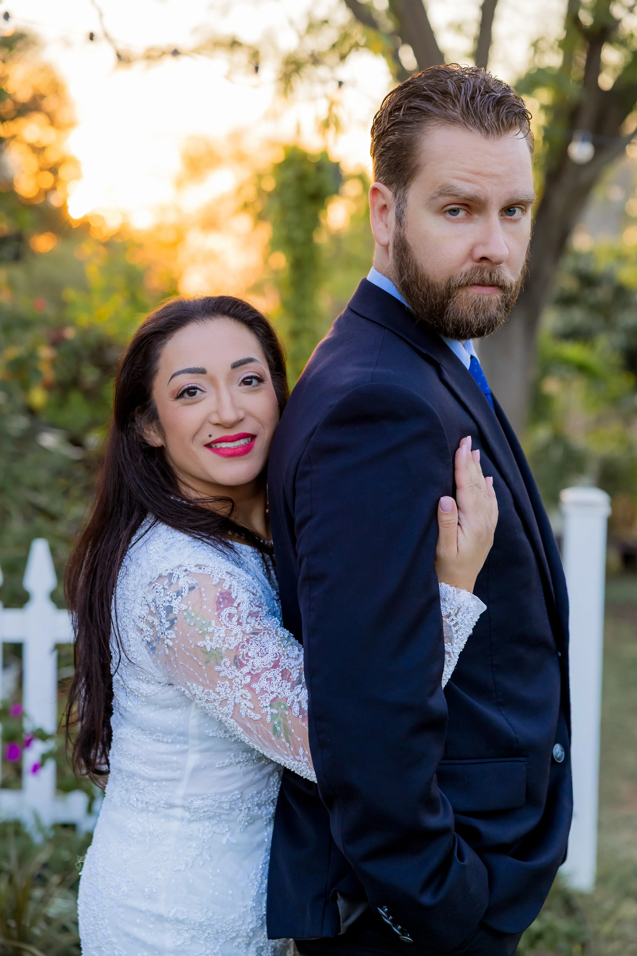 A woman in a white lace dress with long dark hair and red lipstick embracing a man in a navy suit outdoors at sunset, with trees and a white picket fence in the background.