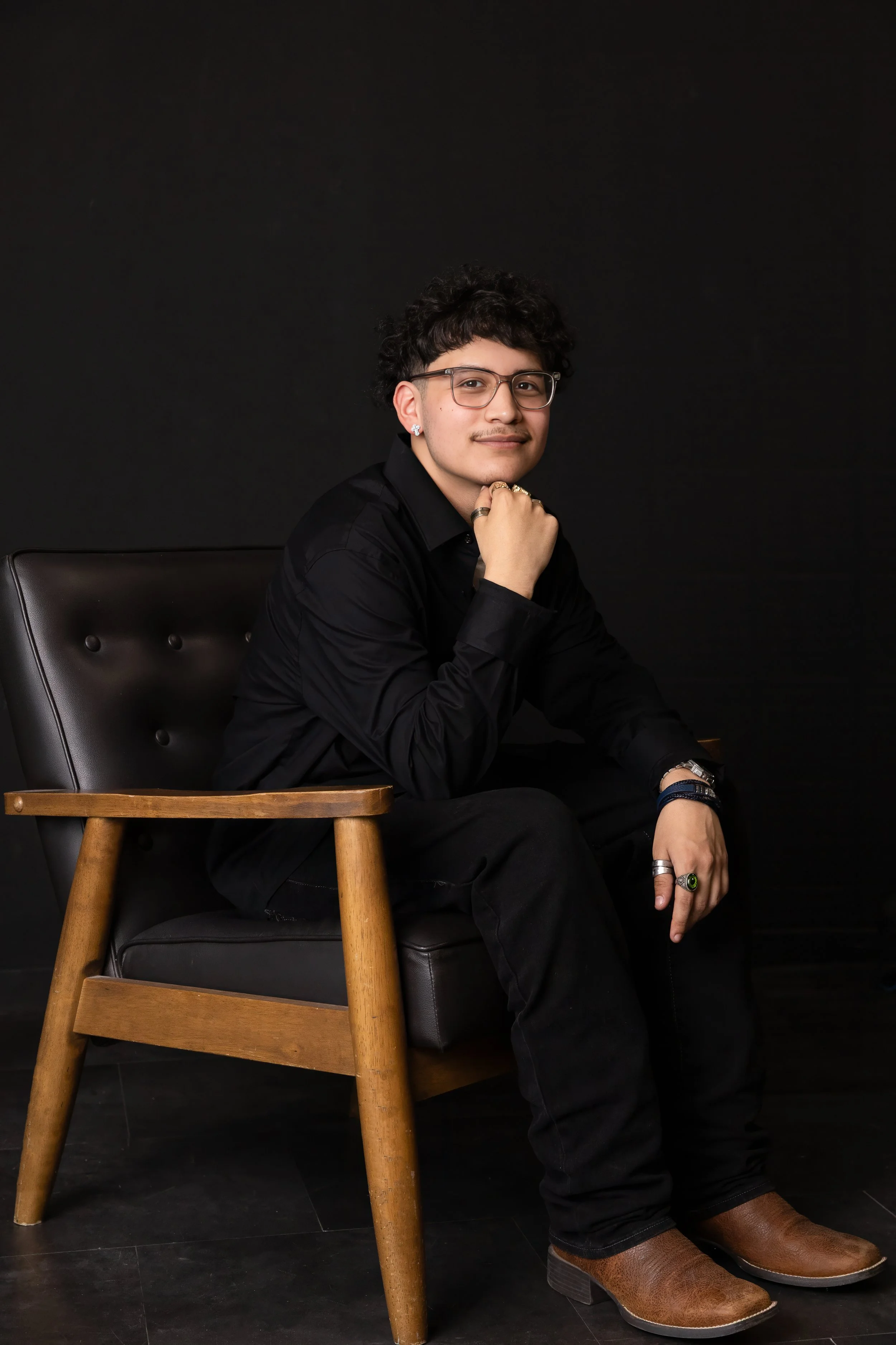 A young man with glasses, earrings, and rings sitting on a black leather armchair with a wooden side table, against a black background, posing with his right hand resting under his chin and smiling.