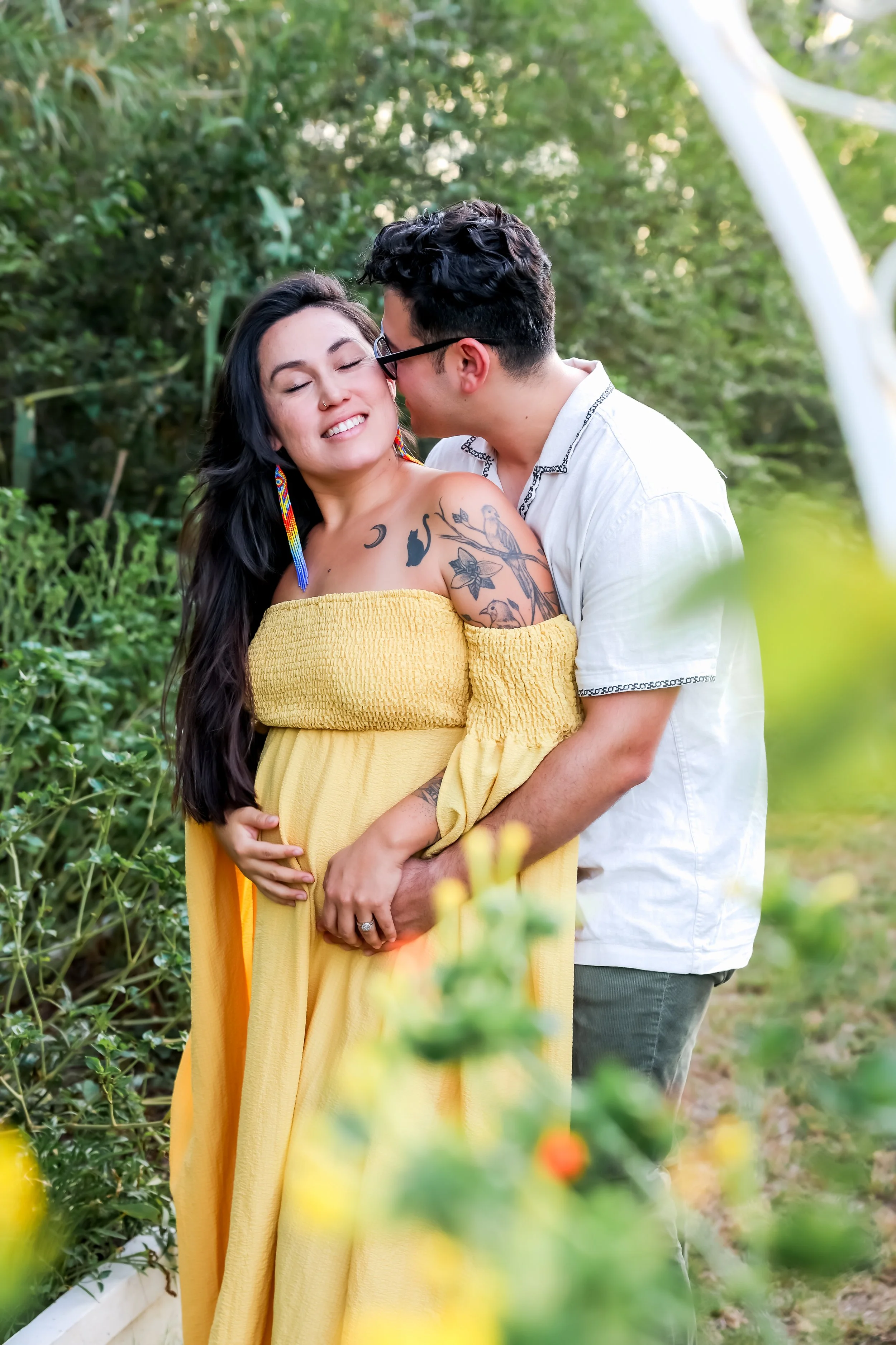 A pregnant woman in a yellow dress is being embraced by a smiling man in a white shirt, outdoors in a lush green setting.