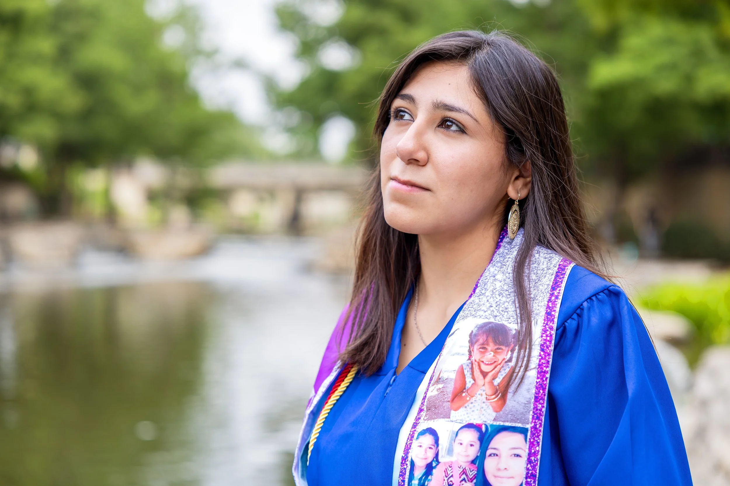 Young woman in graduation gown and honor cords, outdoors near a river, wearing a stole with photos of children, greenery in background.