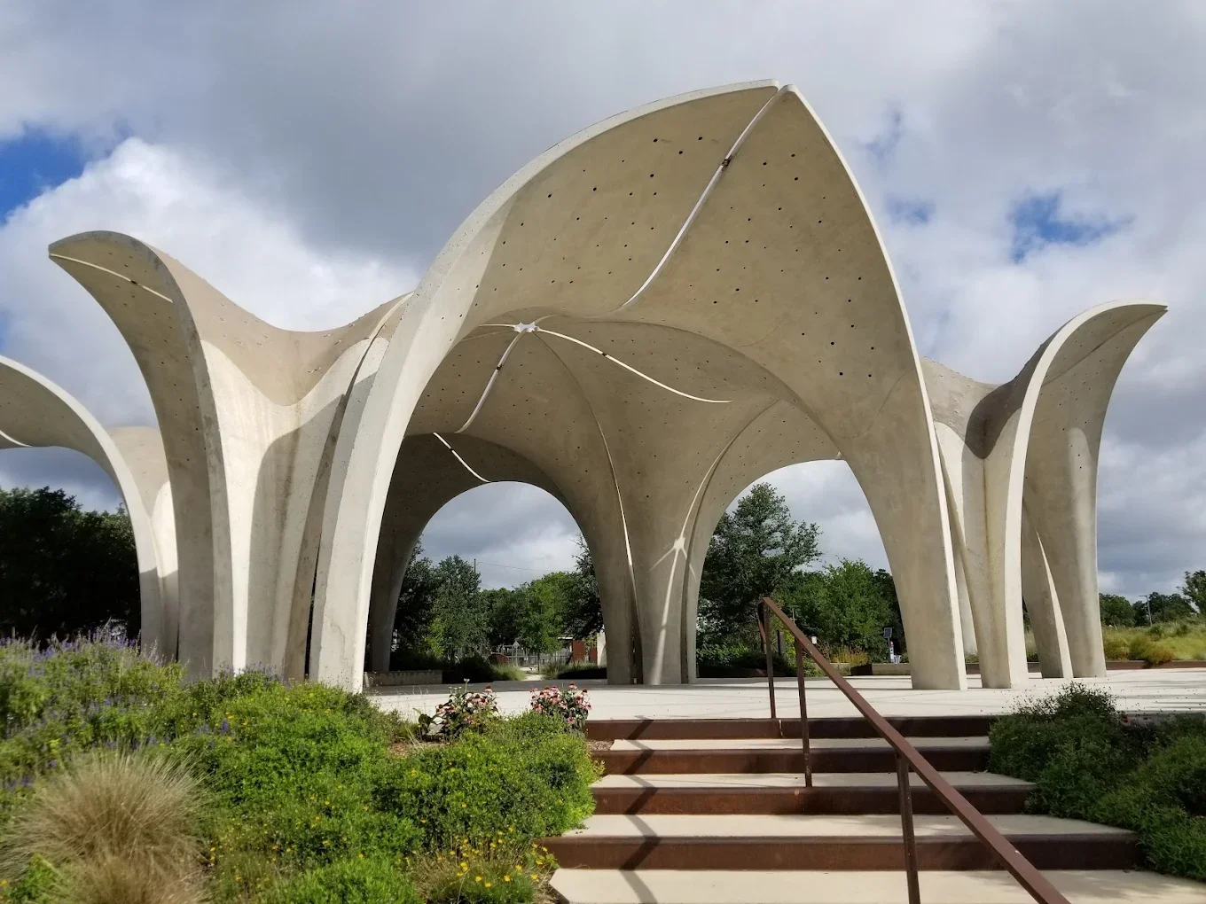 Concrete pavilion with petal-shaped arches and a staircase leading to it, surrounded by greenery and trees under a partly cloudy sky.