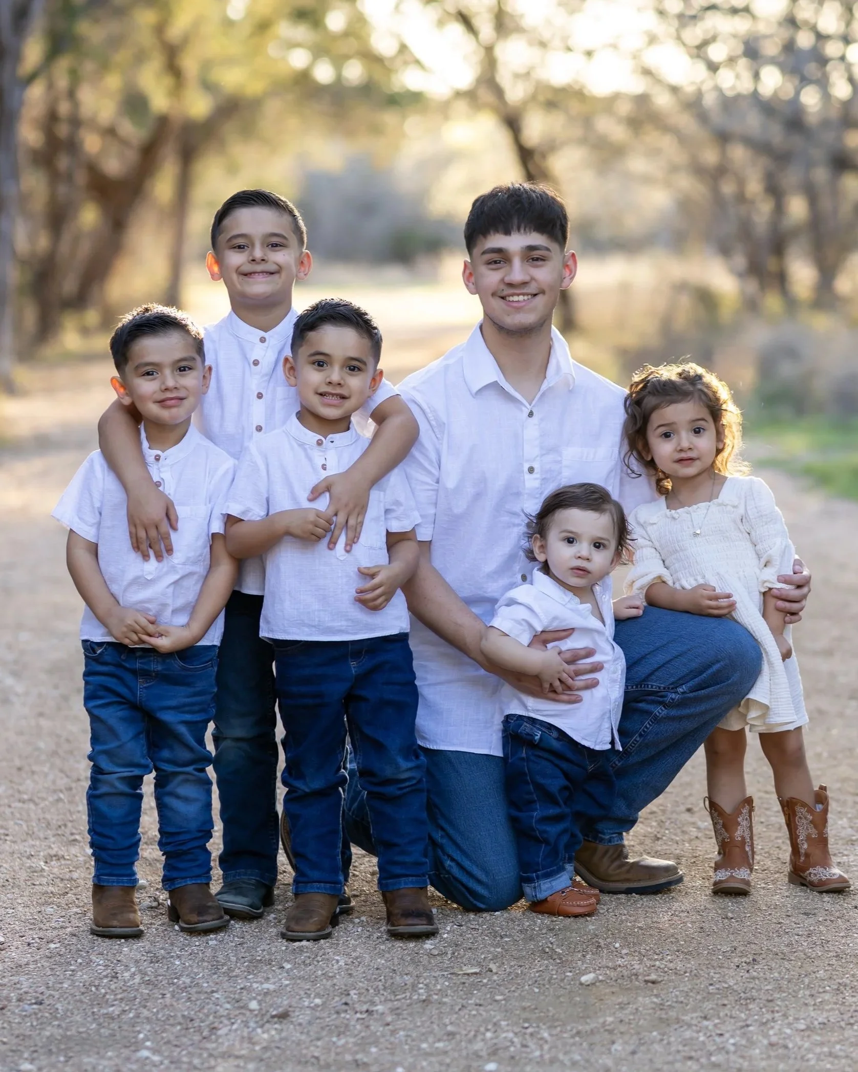 A family of seven, including five children, posing outdoors in a park during daytime with trees in the background. They are dressed in white shirts and blue or beige pants and are smiling at the camera.
