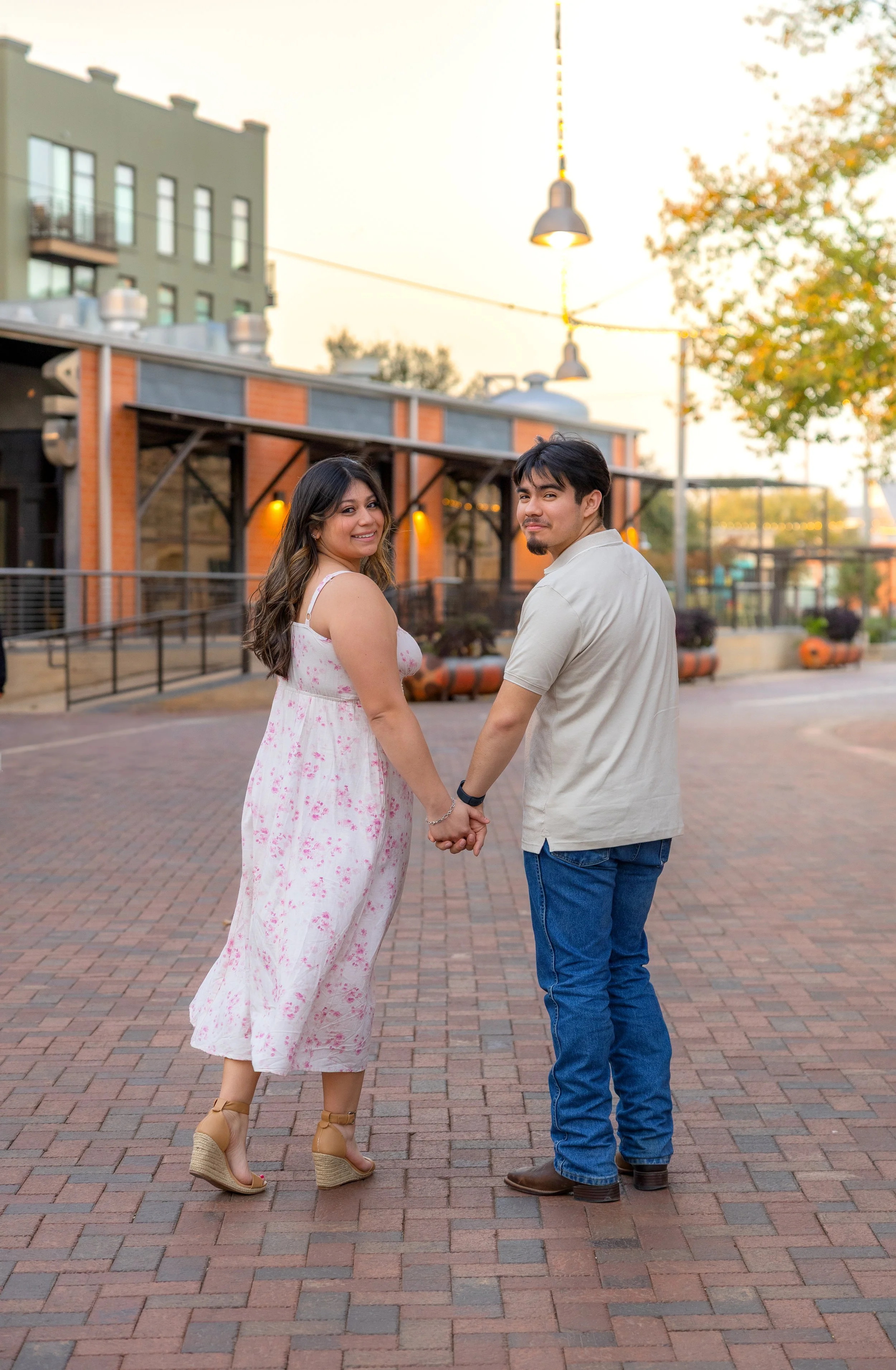 A couple holding hands and smiling at the camera while walking on a brick-paved street at sunset, with buildings and trees in the background.