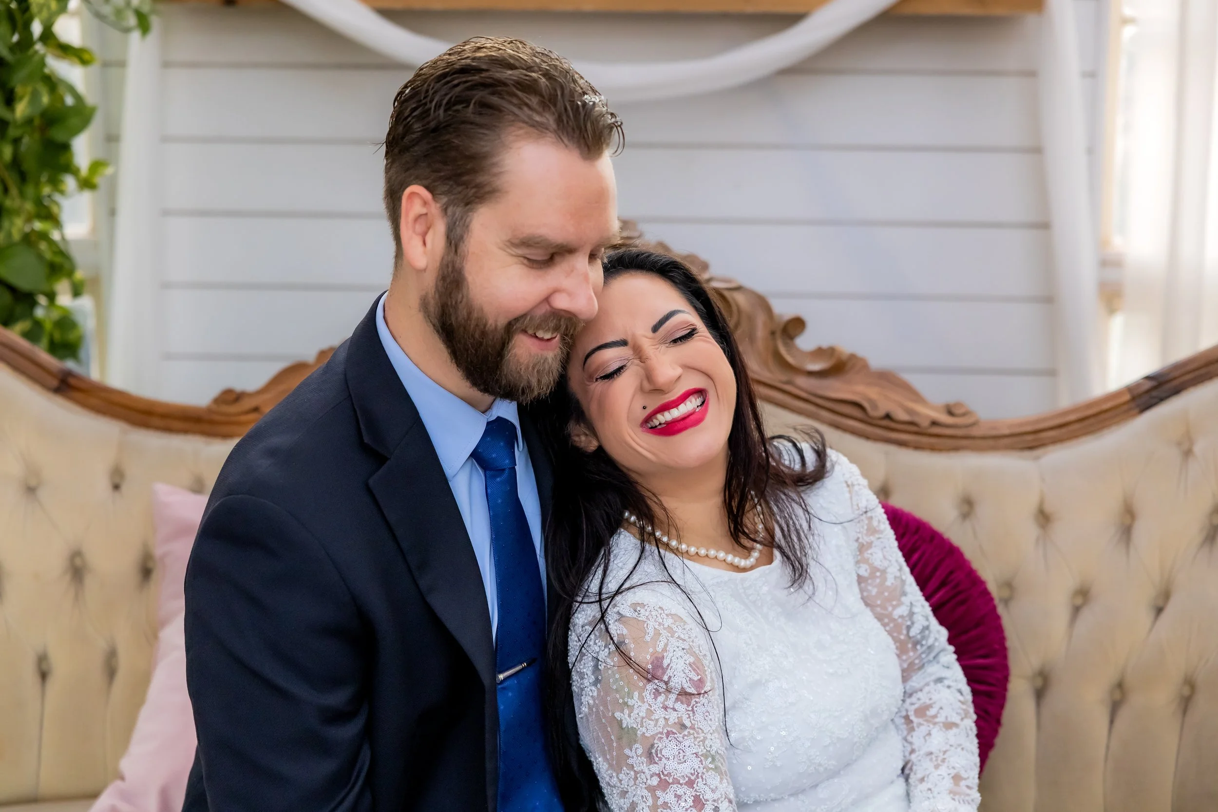 A happy couple, a man with a beard in a suit and a woman in a wedding dress, sharing a joyful moment on a vintage sofa in a bright room.