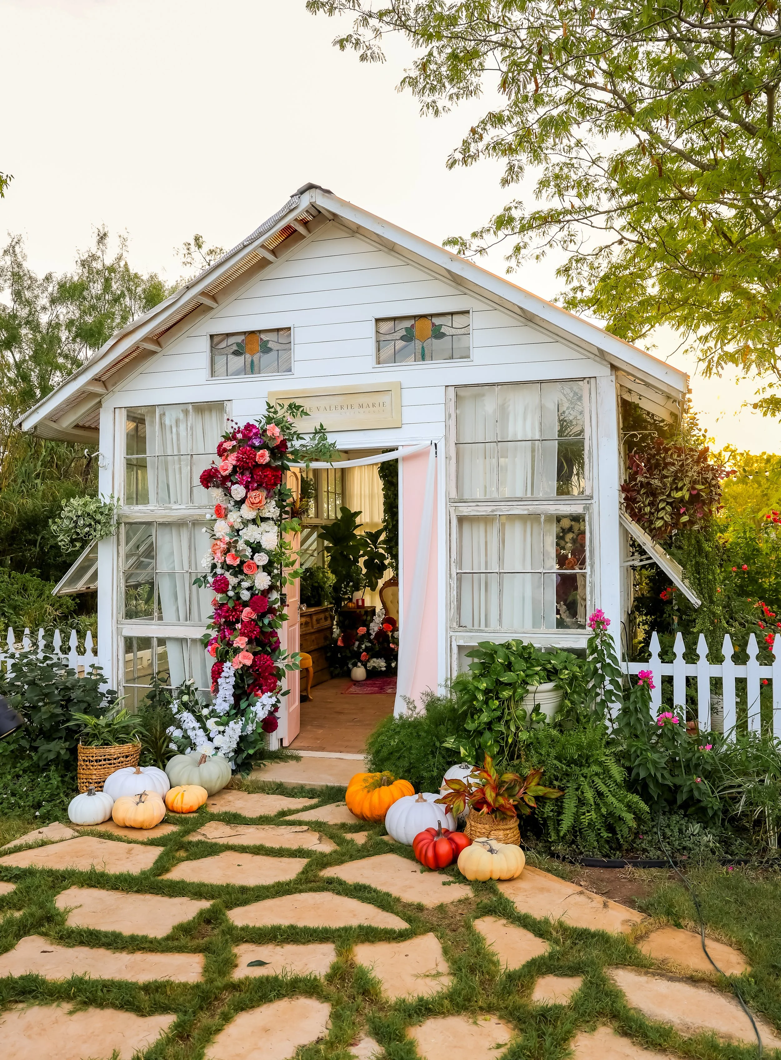 A small white house decorated for a celebration with pumpkins, flowers, and greenery outside the entrance, with a floral arch and a sign above the door that reads 'Valerie Marie'; the scene is taken during sunset.