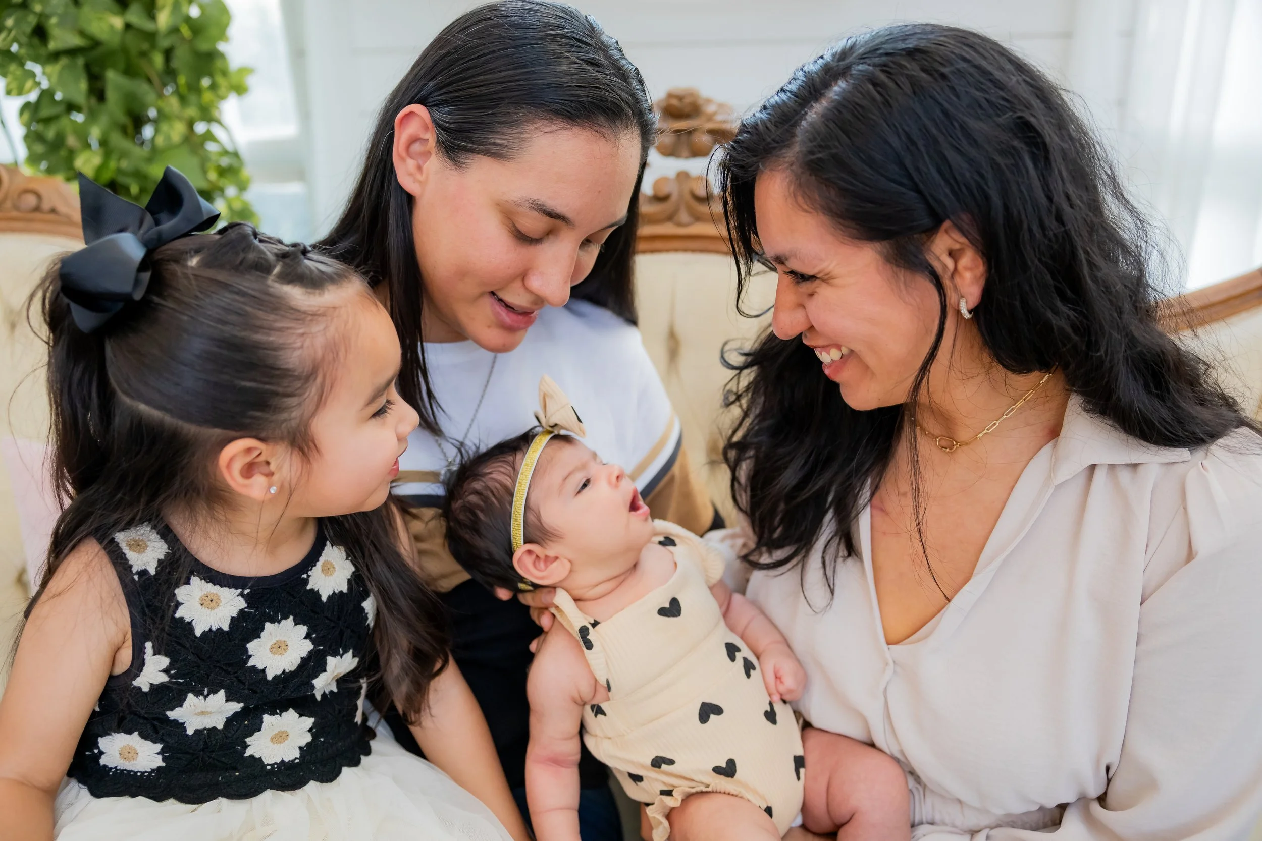 Four women, including a baby, and a young girl, gathered closely with smiles, sharing a sweet moment indoors.