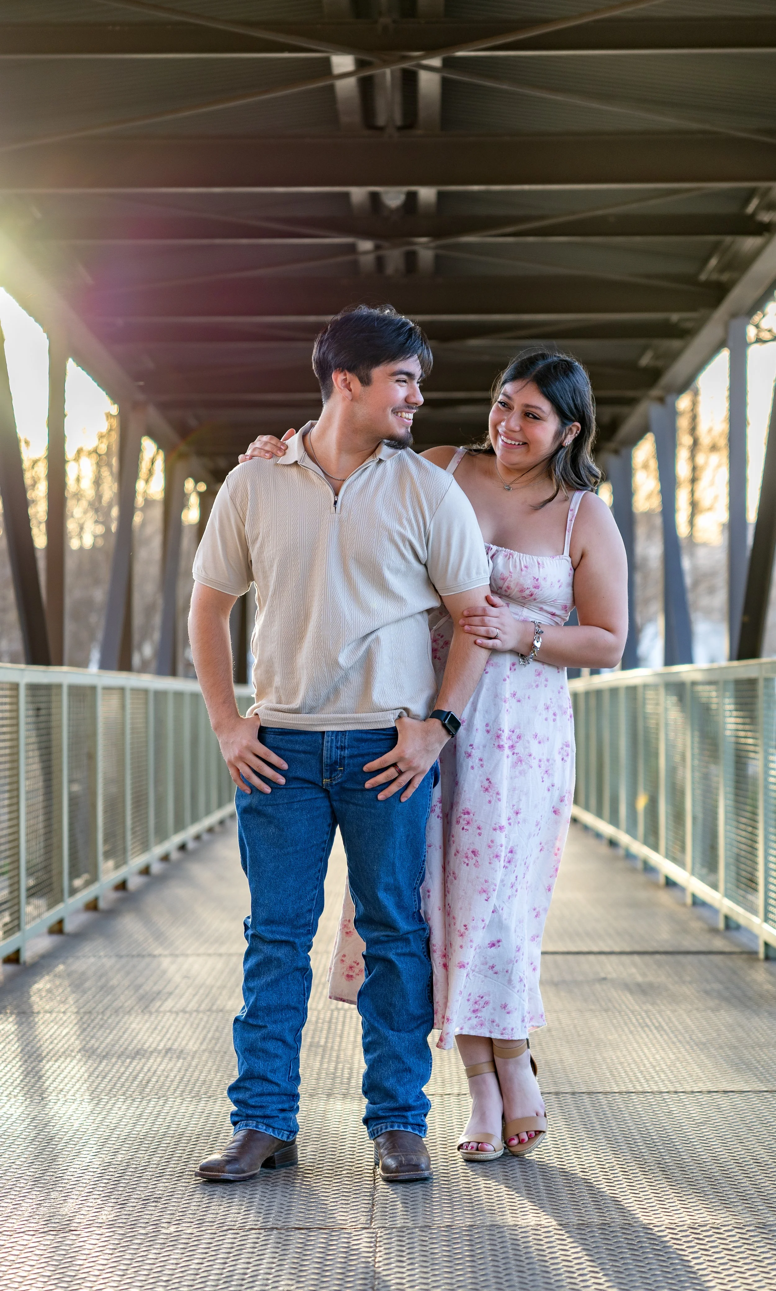 A young couple standing on a metal bridge during sunset, smiling and looking at each other.