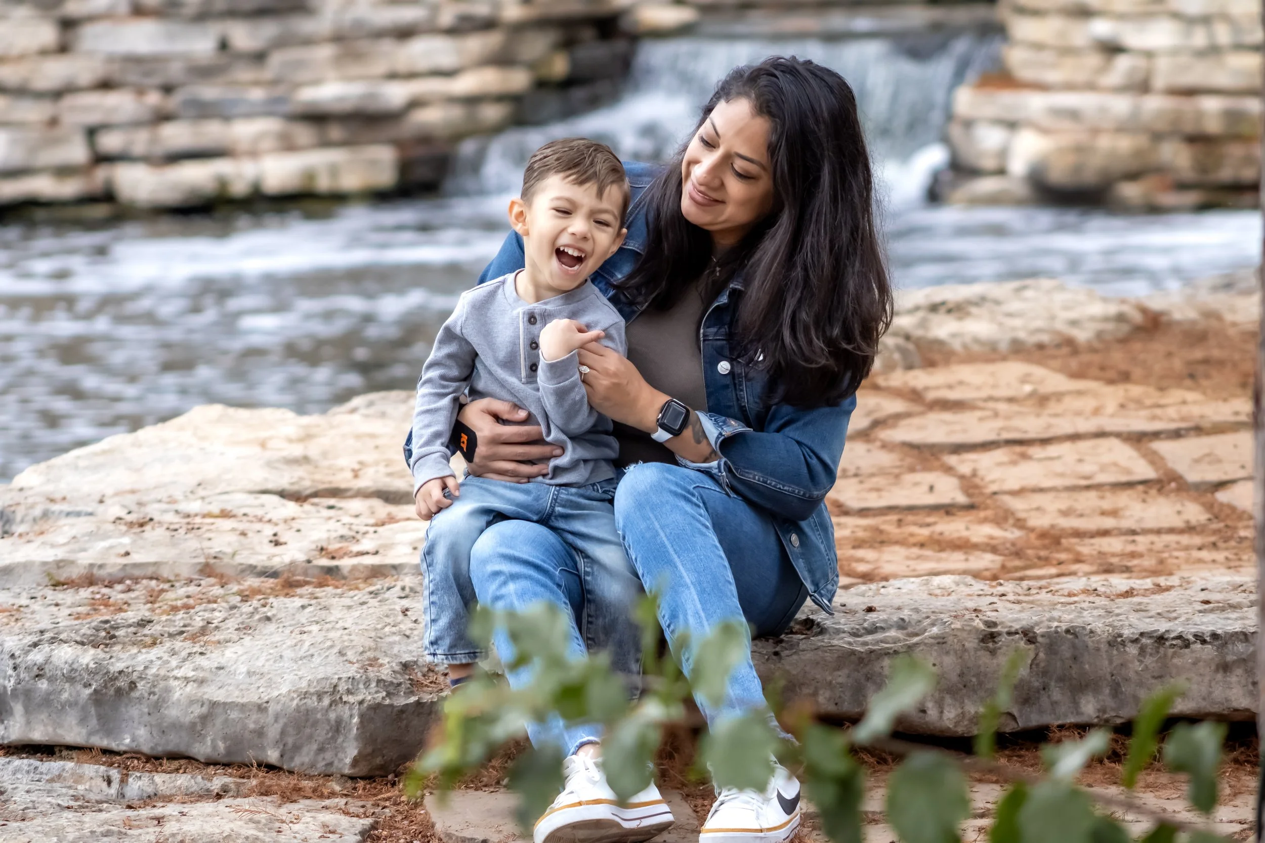 A woman and a young boy sitting on a stone ledge by a river, laughing and enjoying a moment outdoors.