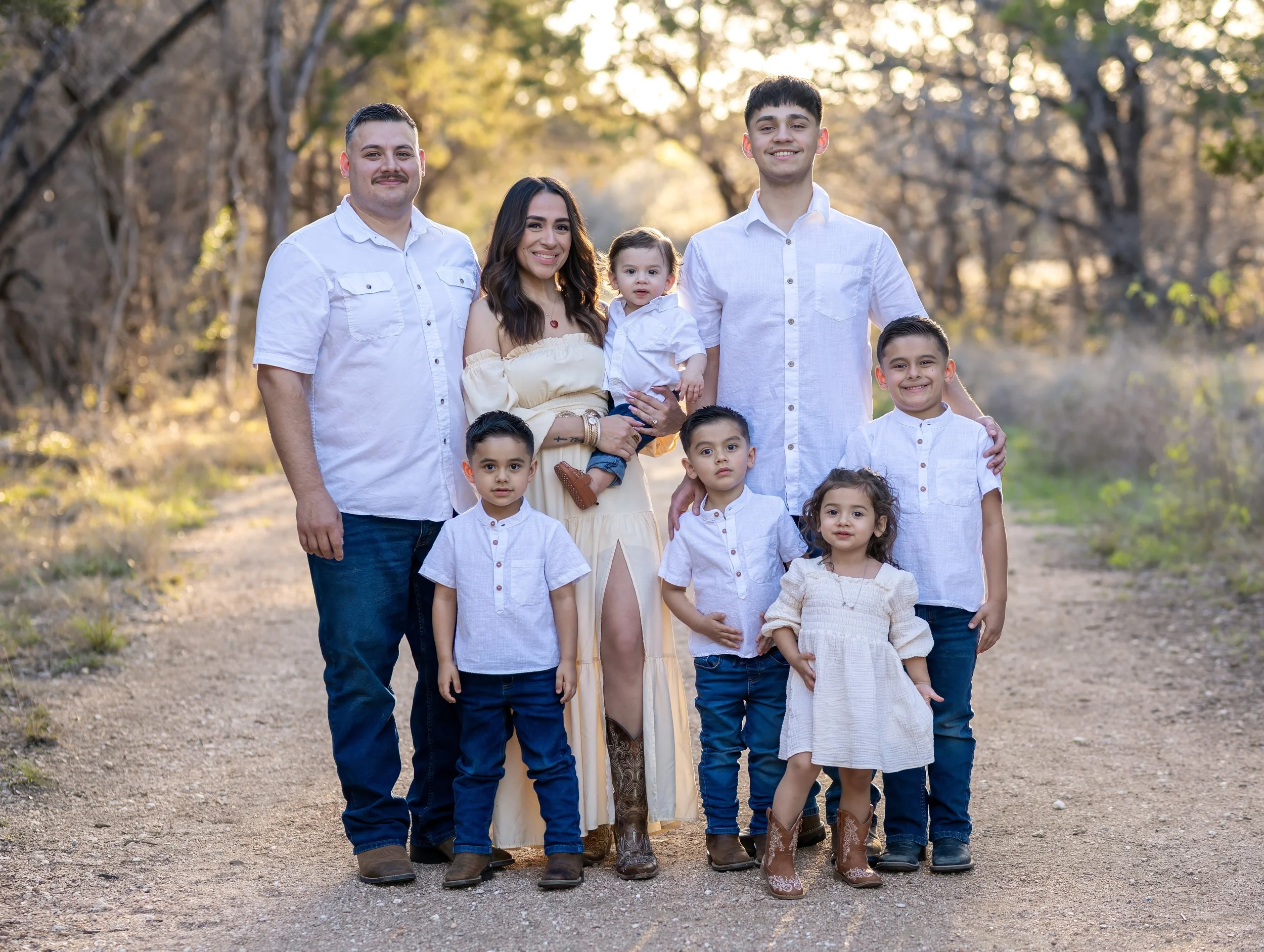 Family of nine posing outdoors in fall, with trees and sunlight in the background. They are dressed in coordinated white and neutral-colored clothing.
