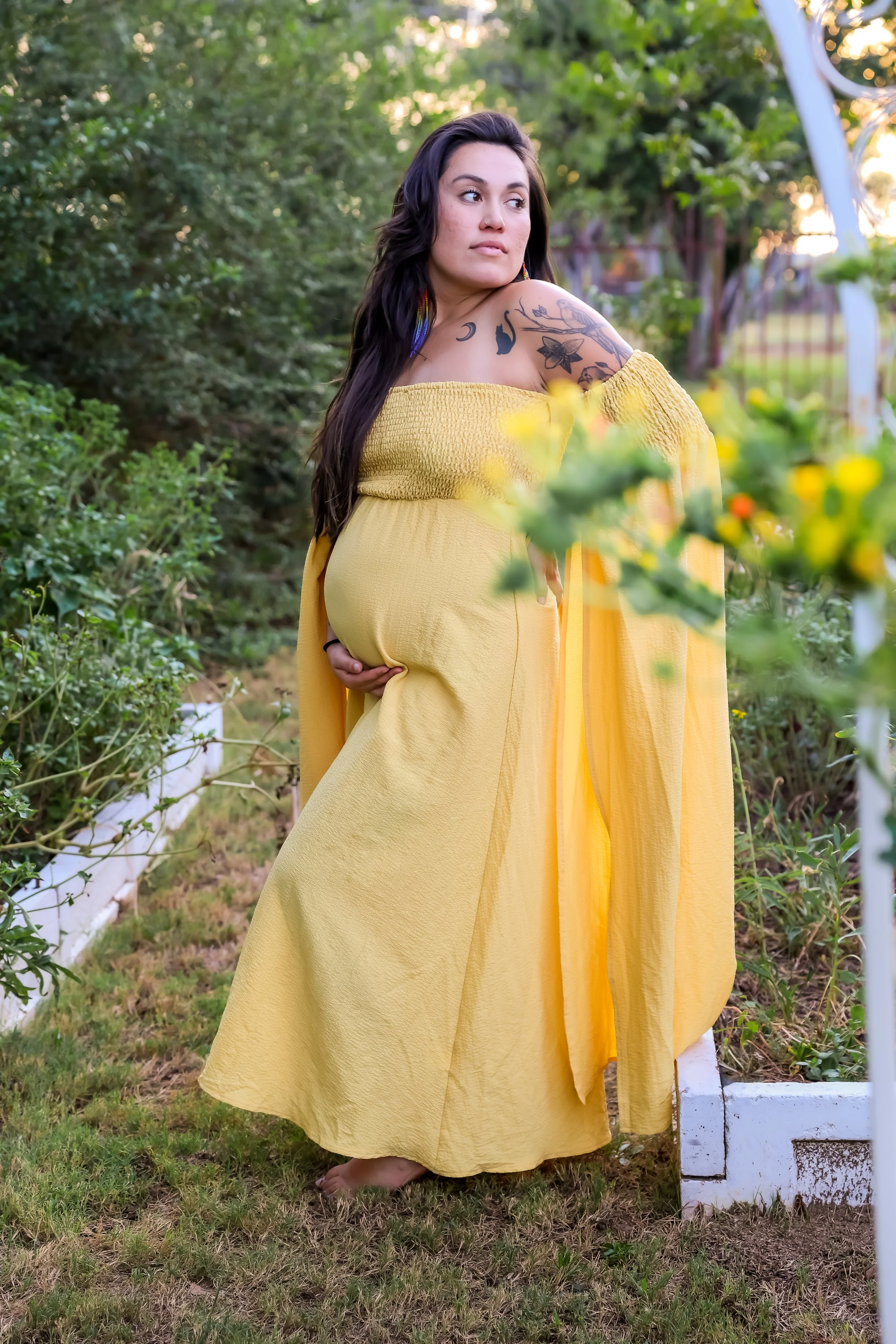 Pregnant woman with long dark hair and tattoos on her arm, wearing a yellow off-shoulder dress, standing outdoors in a garden with green plants and trees.