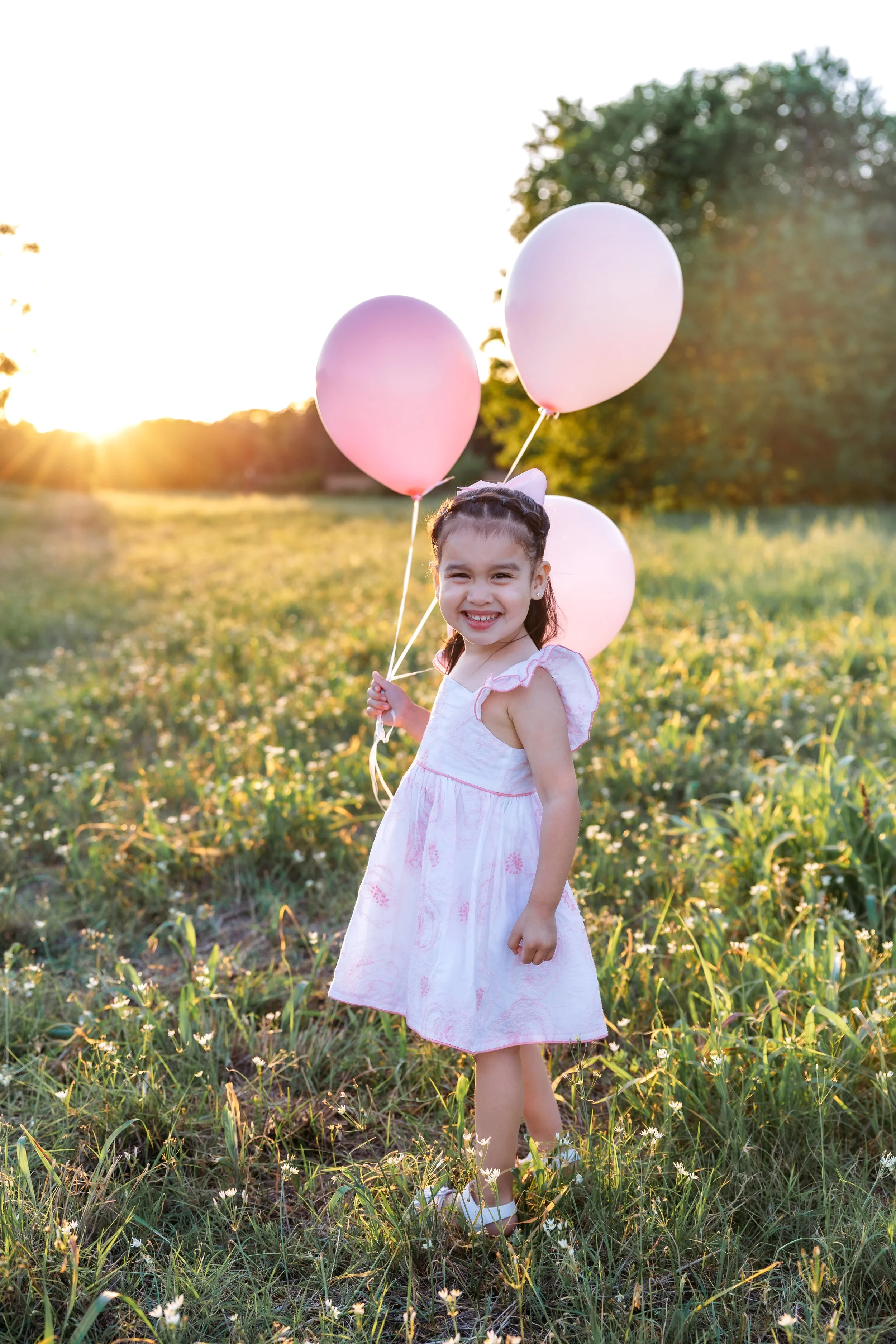 A girl in a pink dress holding three pink and white balloons outdoors at sunset.
