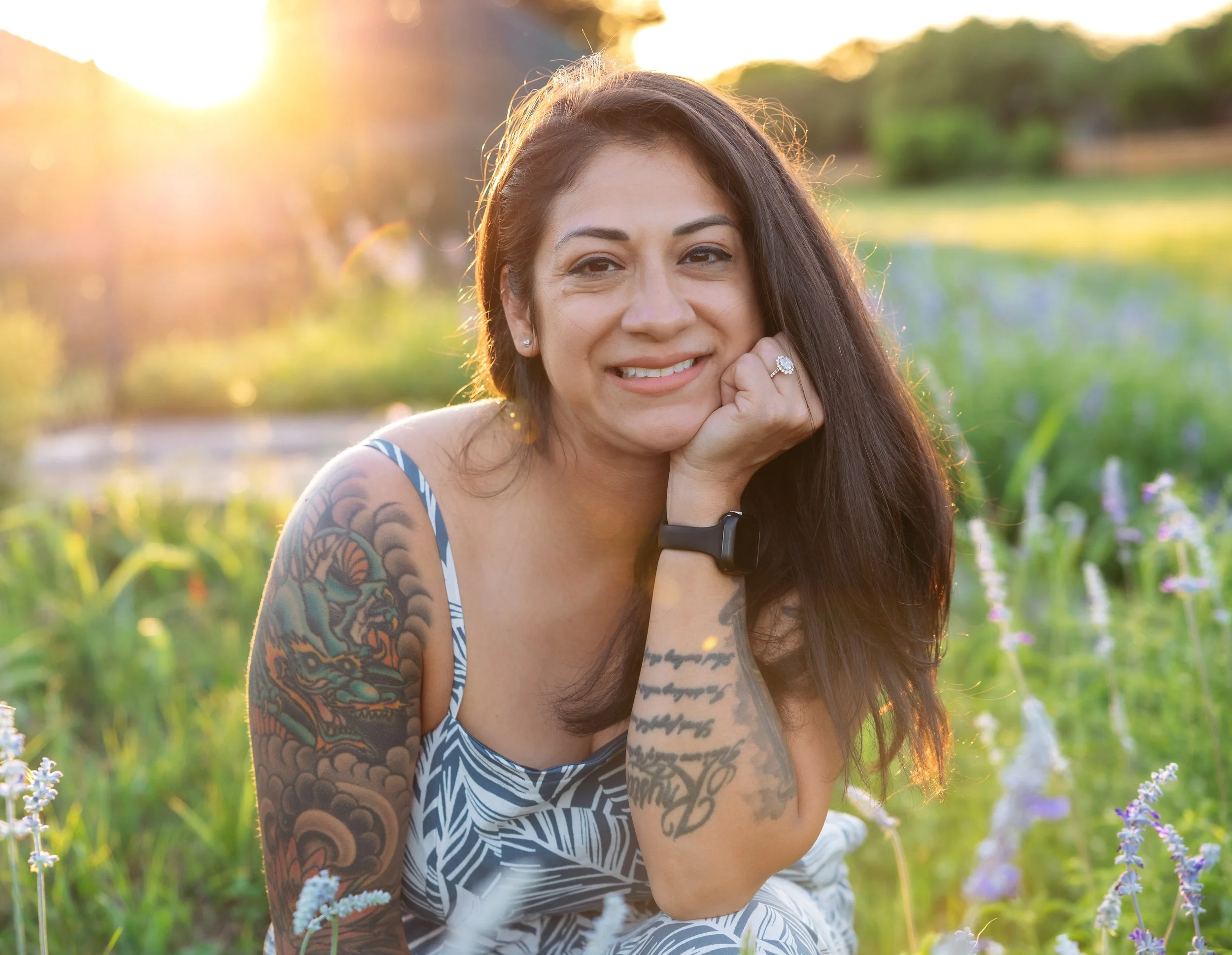 A woman sitting in a field of flowers during sunset, smiling, with long dark hair, tattoos on her arms, wearing a patterned dress, a watch, and a ring.