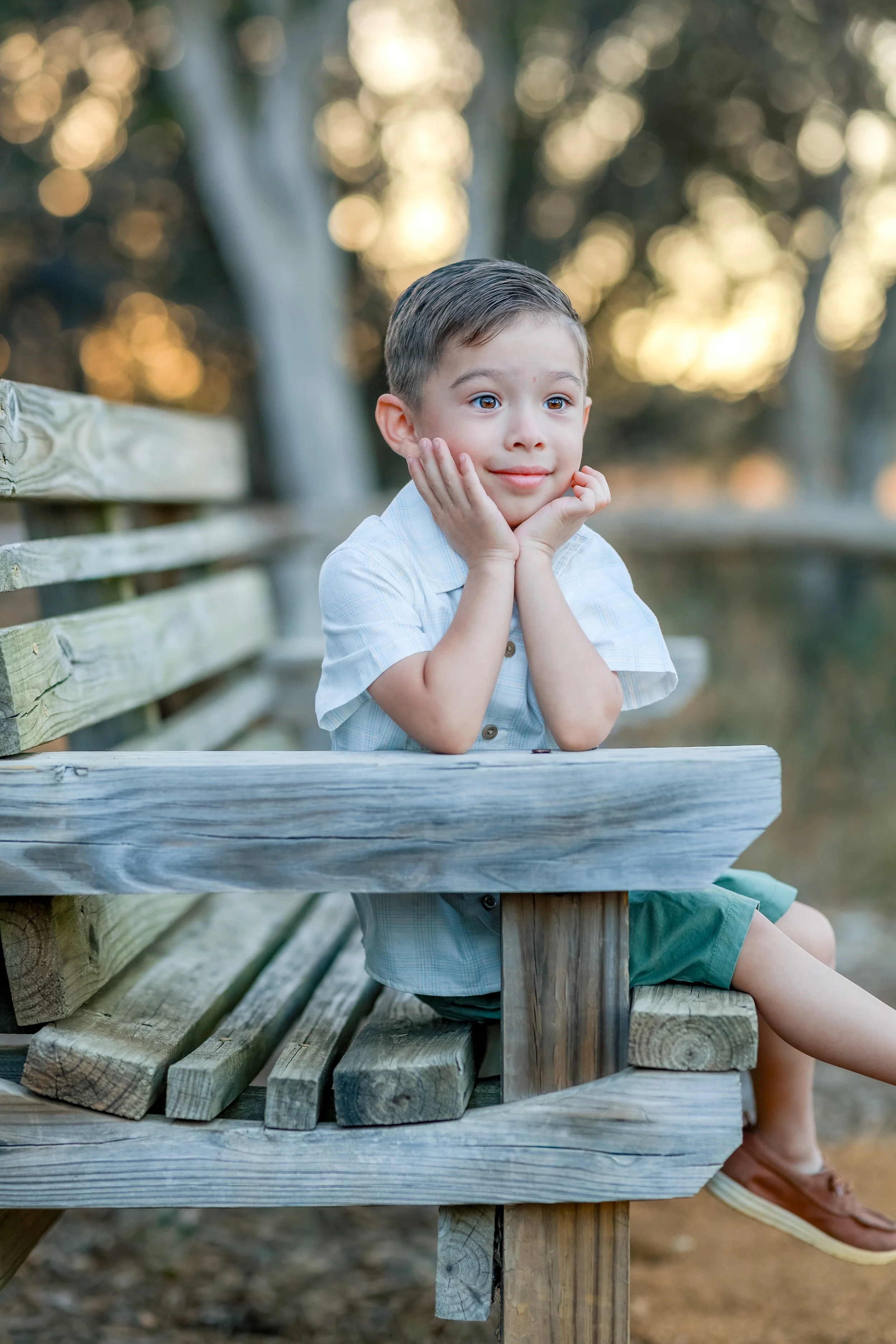 A young boy with short brown hair, sitting on a wooden bench outdoors, resting his chin on his hands, looking thoughtfully into the distance, with trees and soft sunlight in the background.