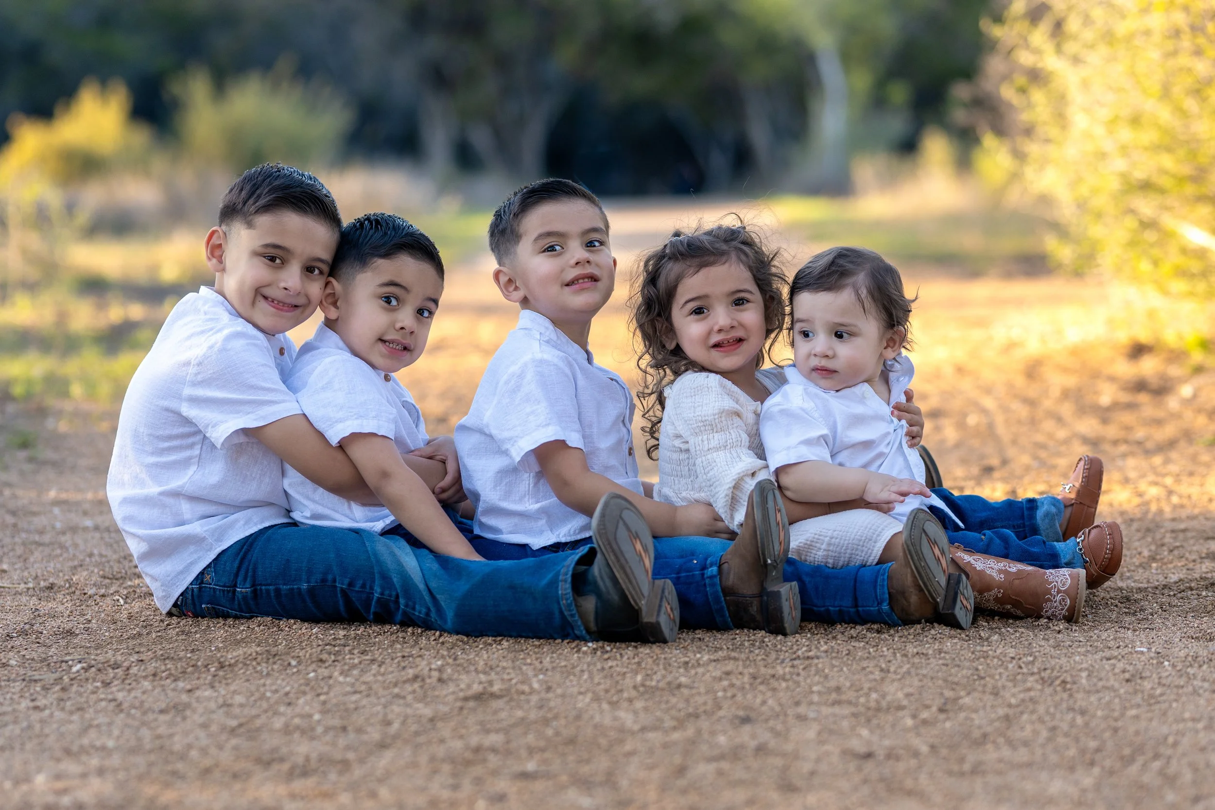 Group of five young children sitting outdoors on dirt path during daytime, wearing casual white shirts and jeans, with trees and sunlight in the background.