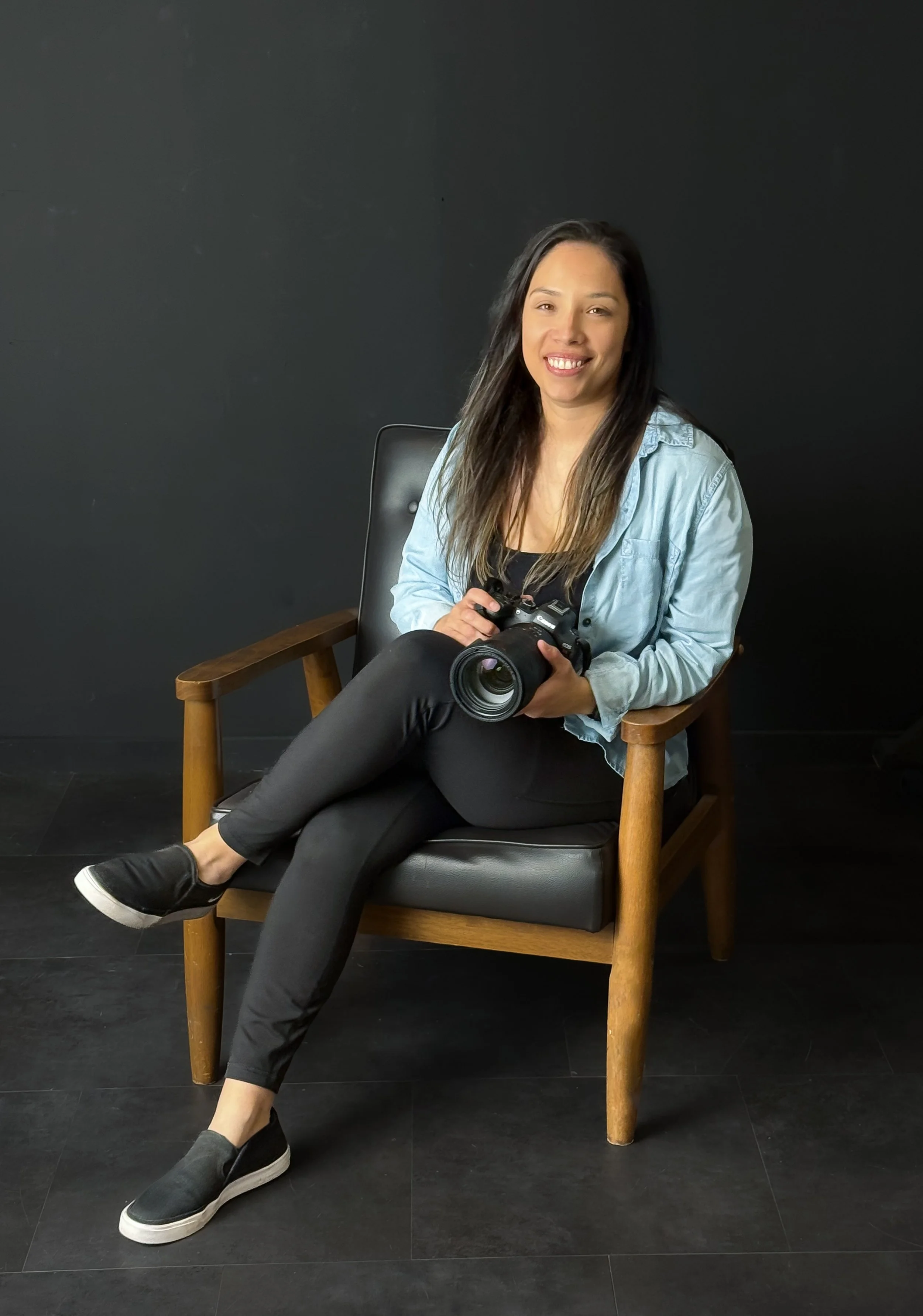 A woman sitting on a wooden chair with a black cushion, holding a camera, smiling, wearing a light blue denim shirt, black pants, and black slip-on shoes, against a black background.