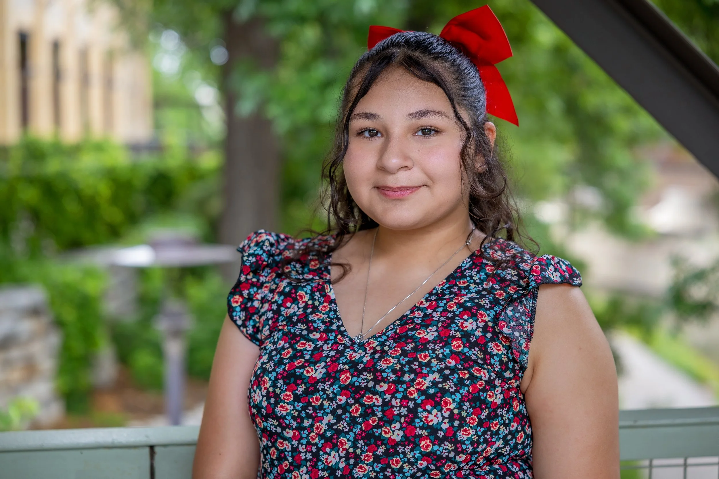 A young girl with curly dark hair wearing a red bow and a floral dress, smiling outdoors with greenery in the background.