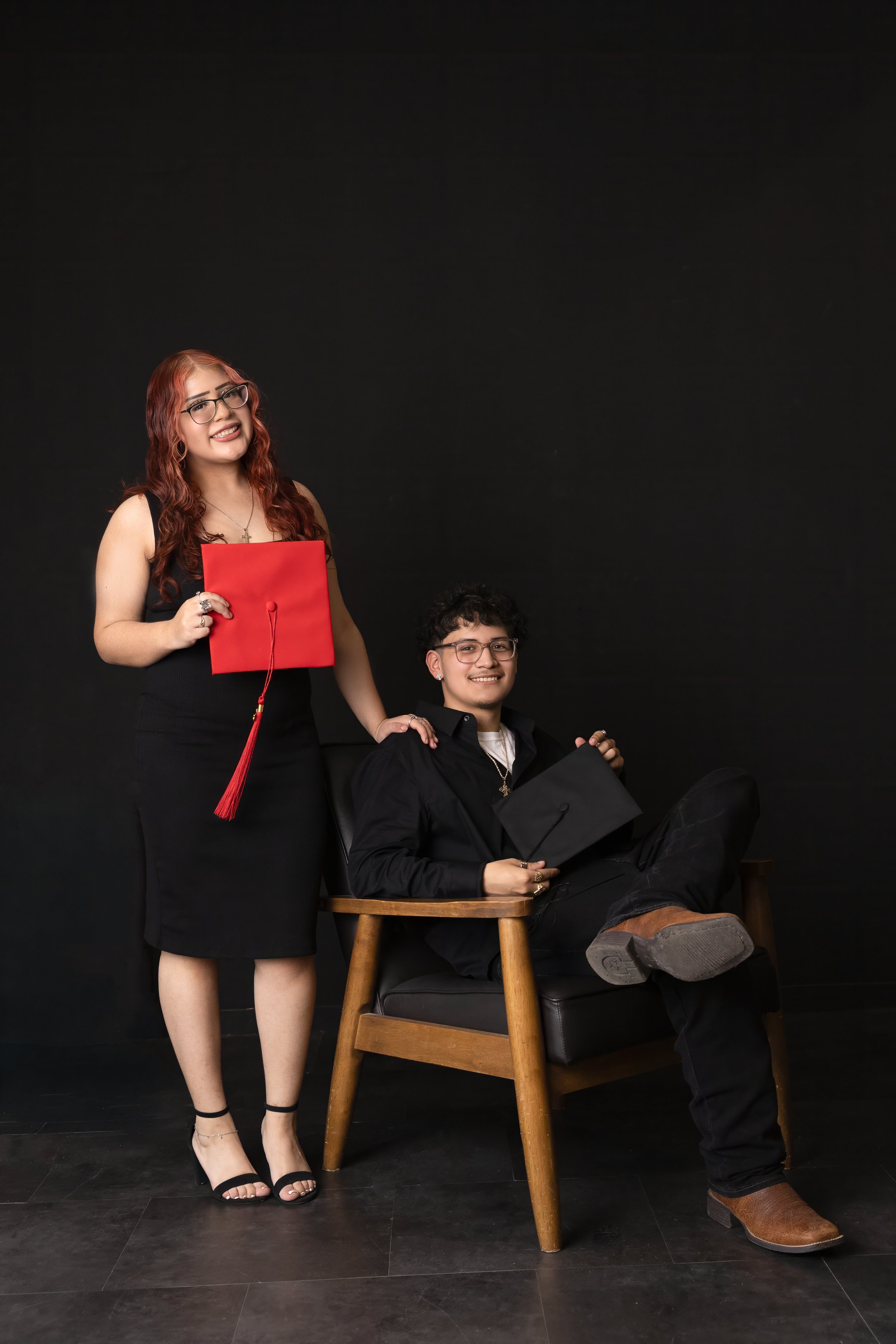 A young man sitting in a chair holding a diploma, with a young woman standing beside him, both dressed in semi-formal attire for a graduation photo against a black background.