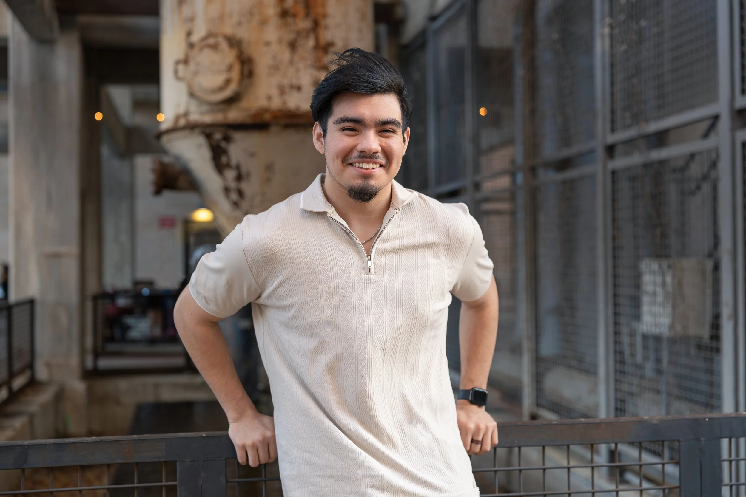 A young man with dark hair, a goatee, and a light beige polo shirt, smiling and leaning on a metal railing outdoors.