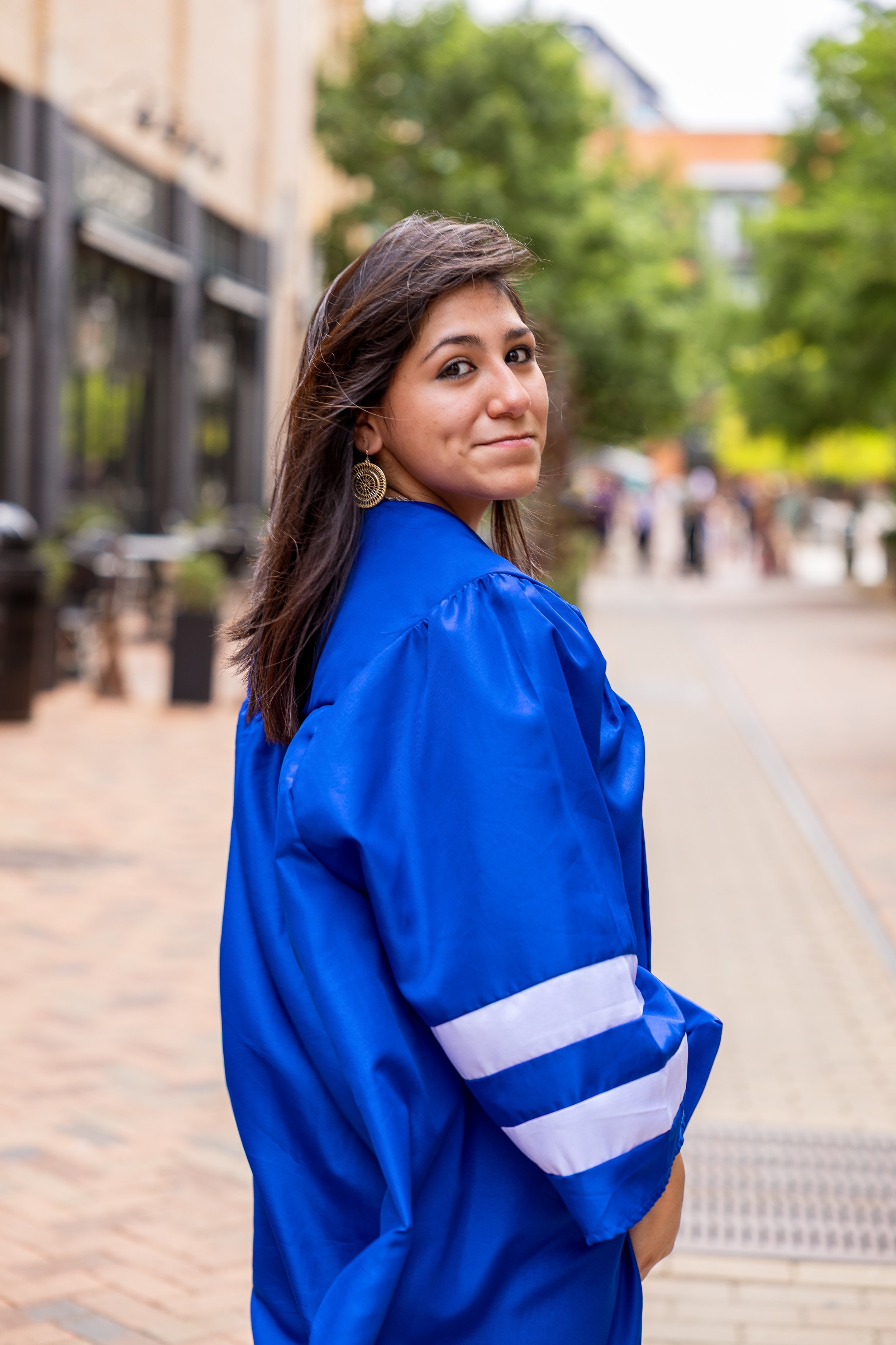 A young woman in a blue graduation gown standing outdoors on a city street with trees and buildings in the background.