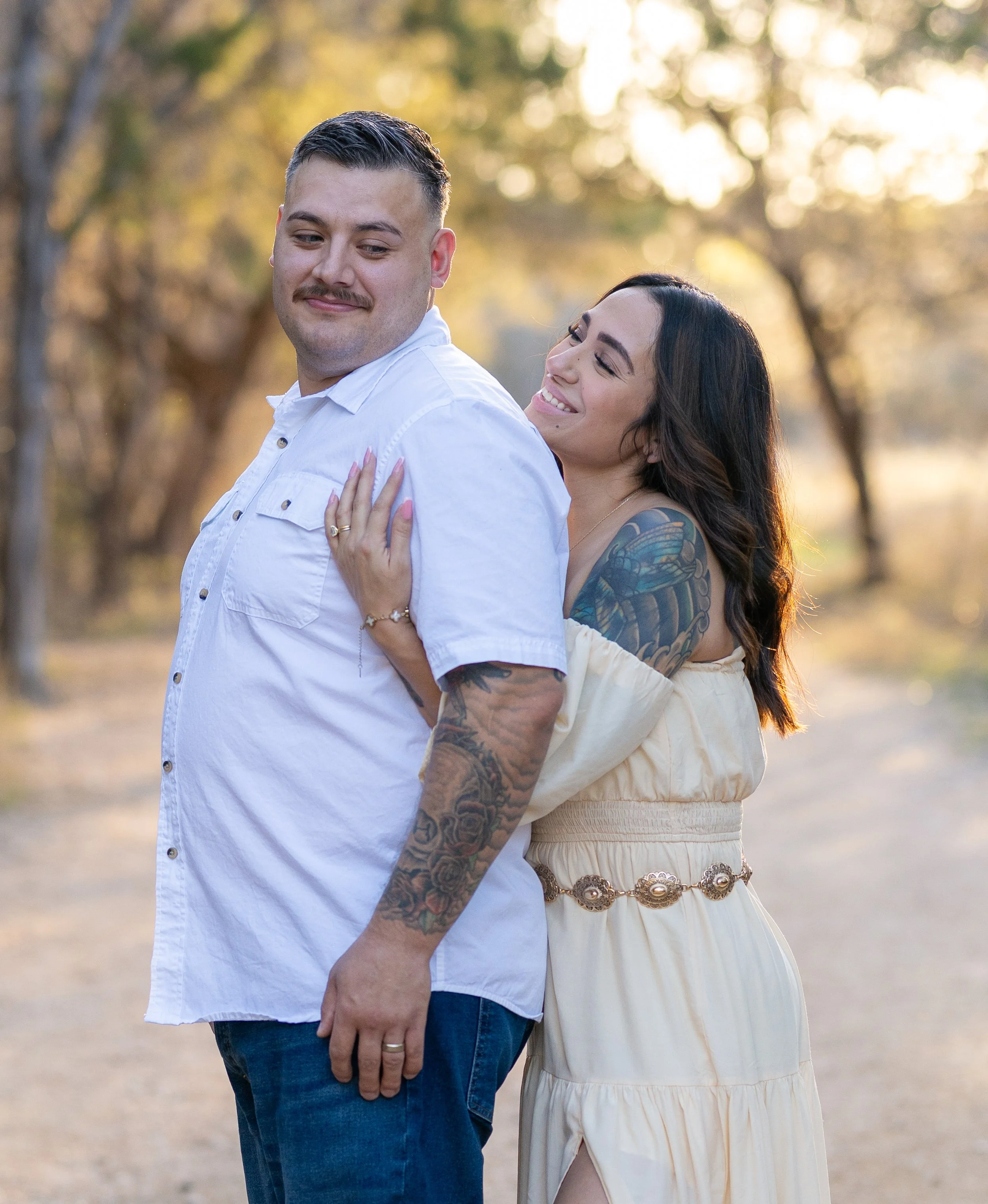 A couple standing outdoors on a dirt path during sunset, with trees in the background. The woman is smiling and embracing the man from behind, who has a neutral expression.