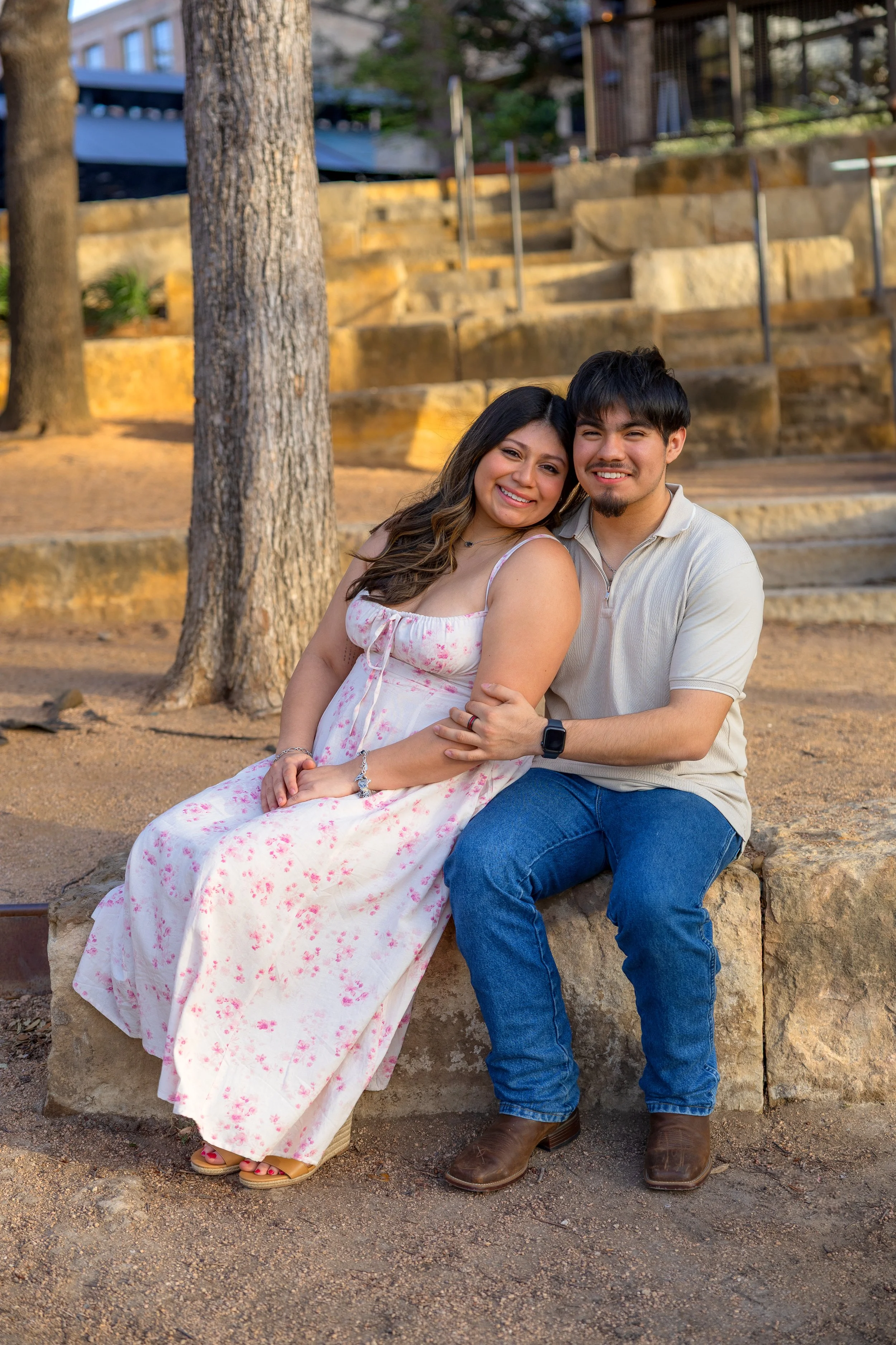 A smiling woman and man sitting close together outdoors, with trees and stone steps in the background.