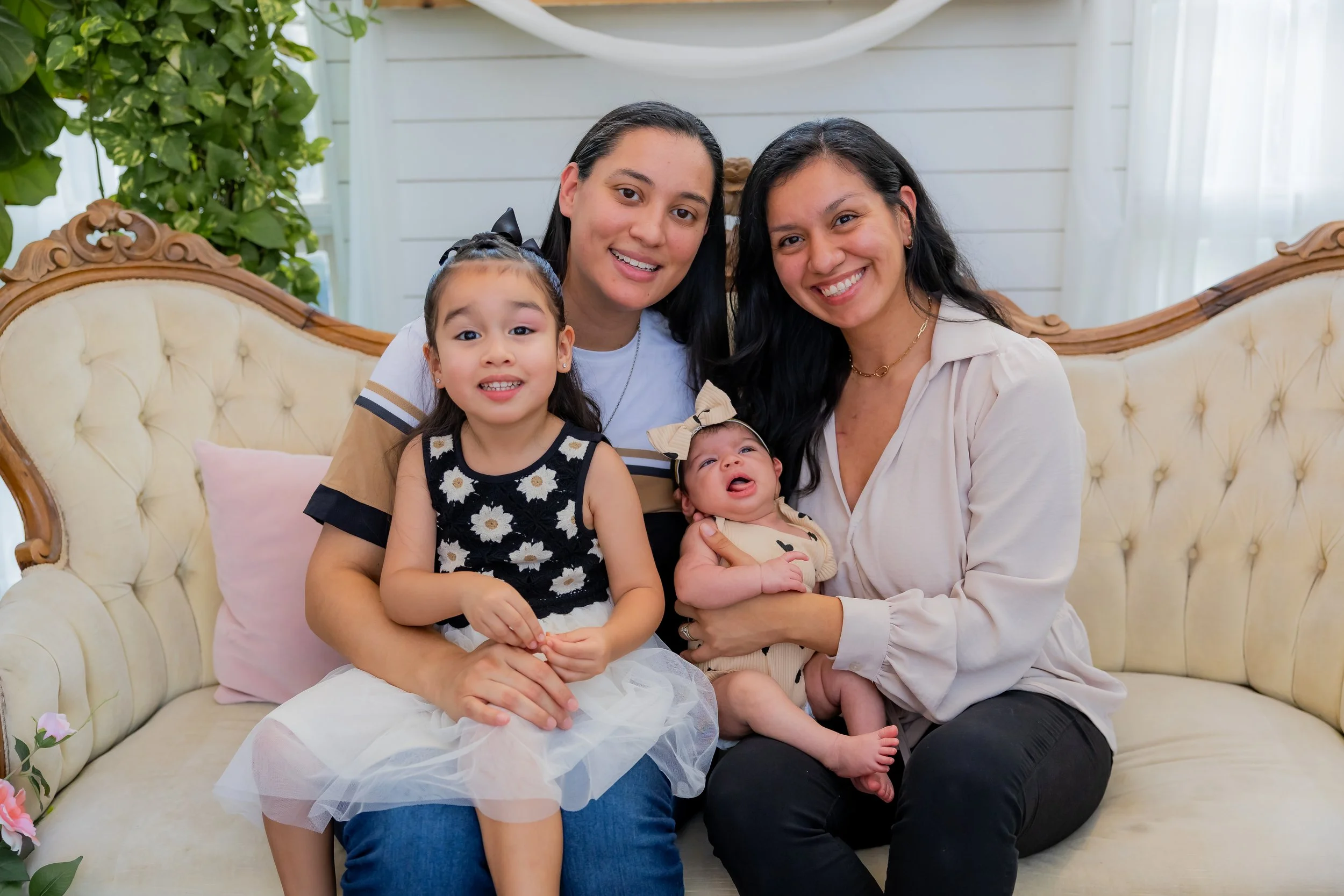 Four women, two children and an infant, sitting on a vintage cream sofa in a bright room with white walls and large windows. They are smiling and posing for the photo.