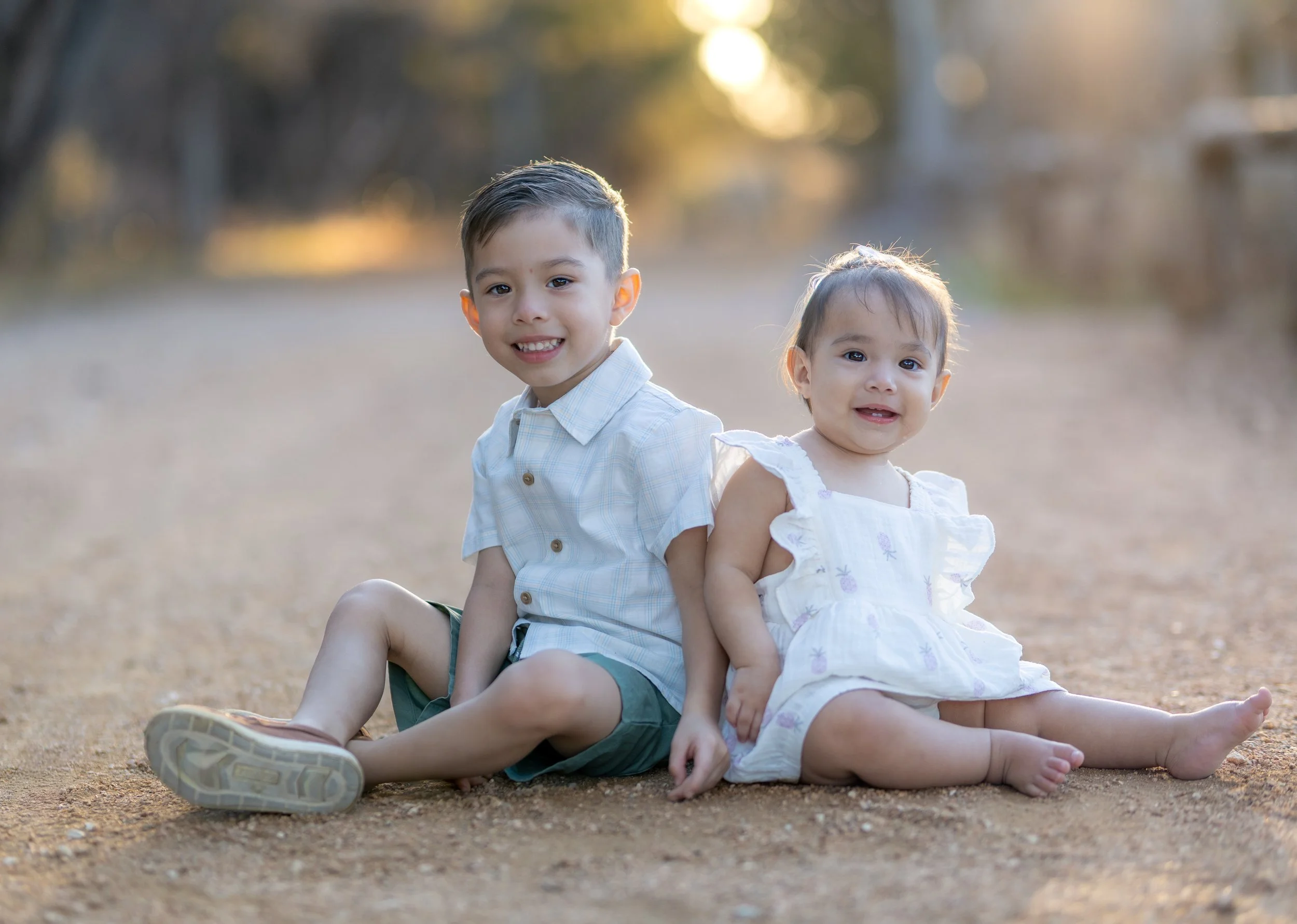 A young boy and a toddler girl sitting on a dirt path outdoors, smiling at the camera, with trees and sunlight in the background.