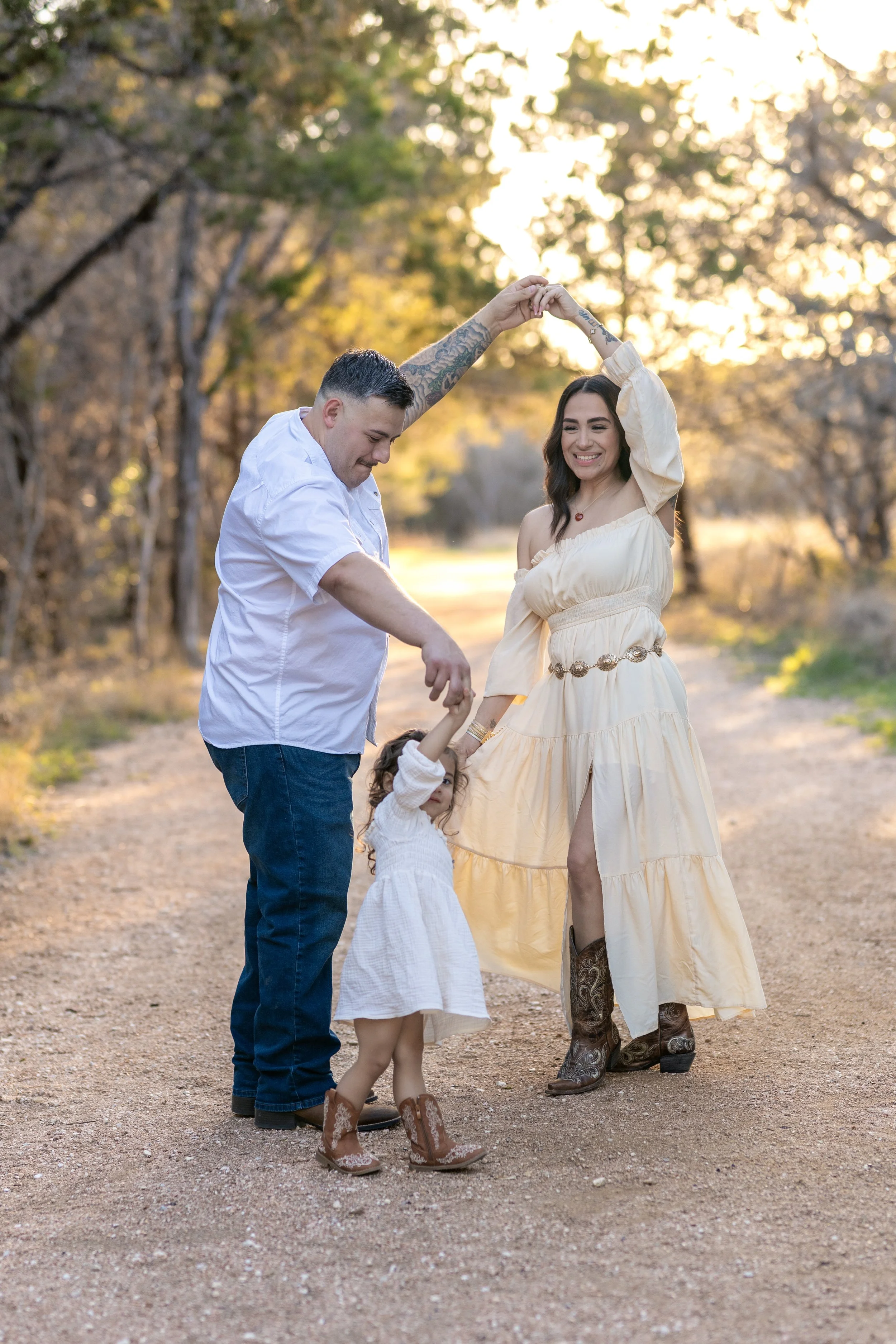 A family of three dancing on a dirt path in a wooded area during sunset. The father, with tattoos and wearing a white shirt and jeans, is holding hands with his young daughter, who is wearing a white dress and cowboy boots. The mother, in a cream-colored dress and cowboy boots, is holding her hand up, smiling.