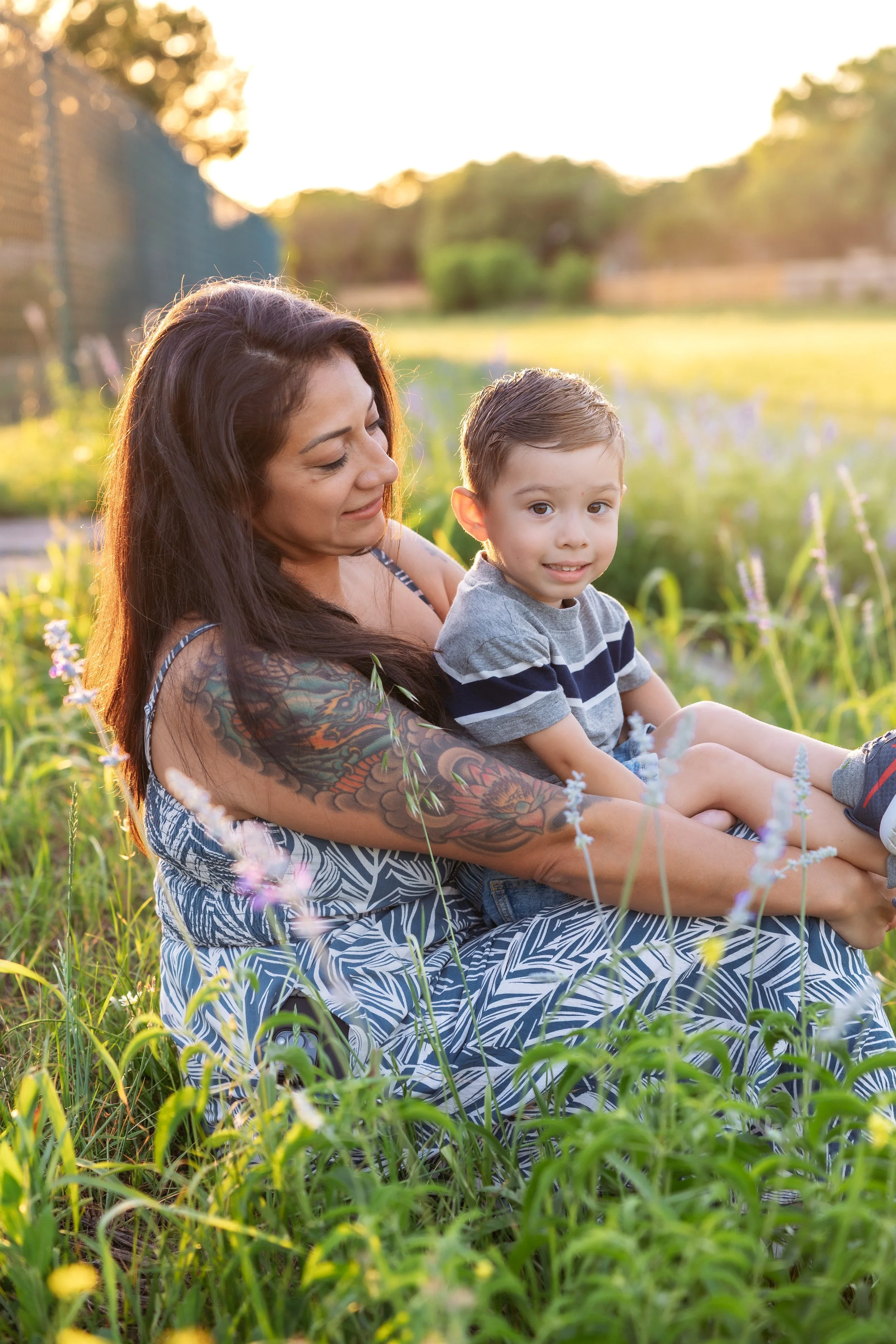 A woman with dark hair and tattoos on her arm sitting in a grassy field with a young boy on her lap. They are outdoors during sunset, surrounded by tall grass and flowers, with the warm sunlight illuminating the scene.
