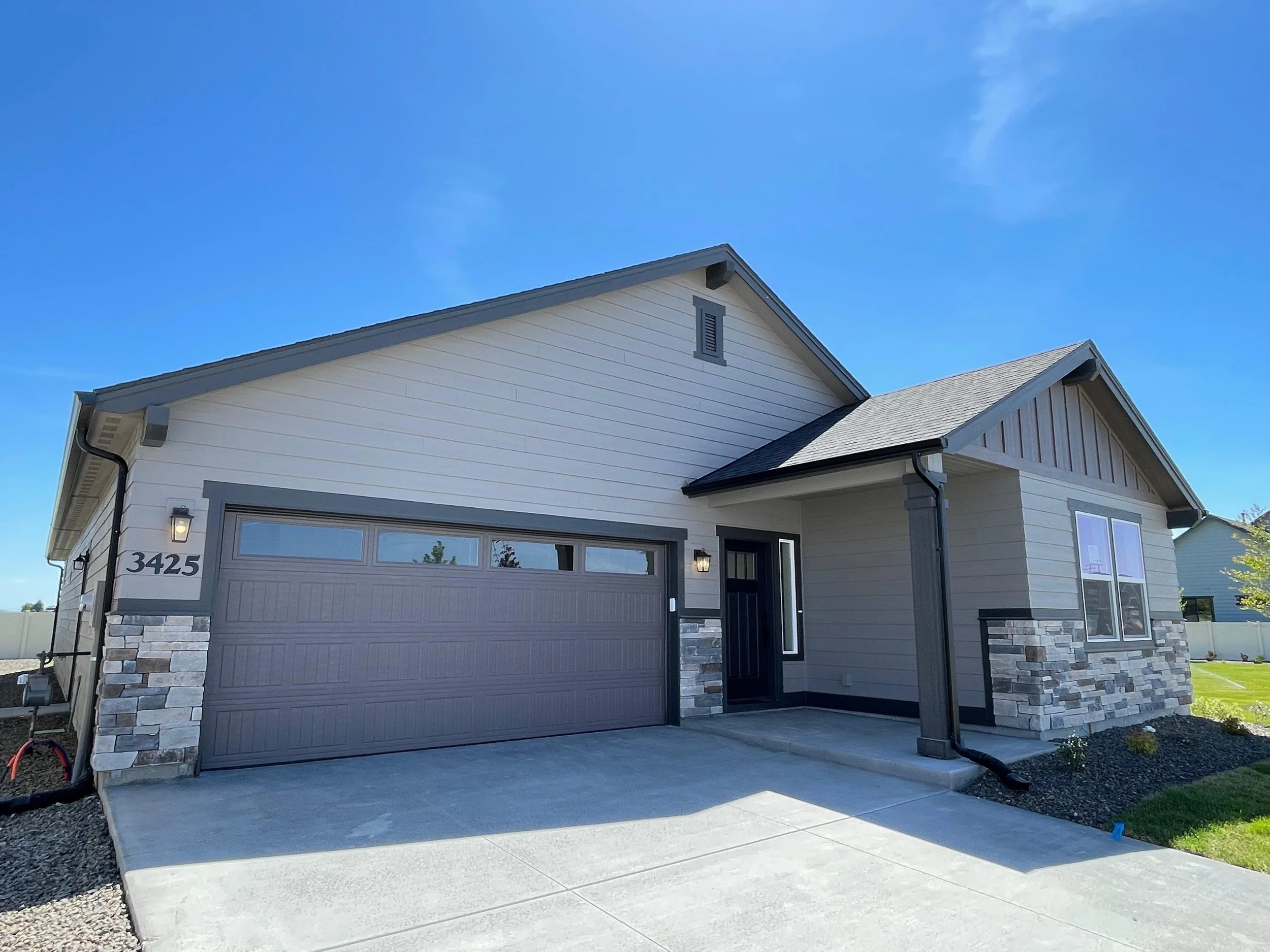 Front of a modern house with a garage, dark gray door, white and gray siding, stone accents, and landscaping, under a clear blue sky.