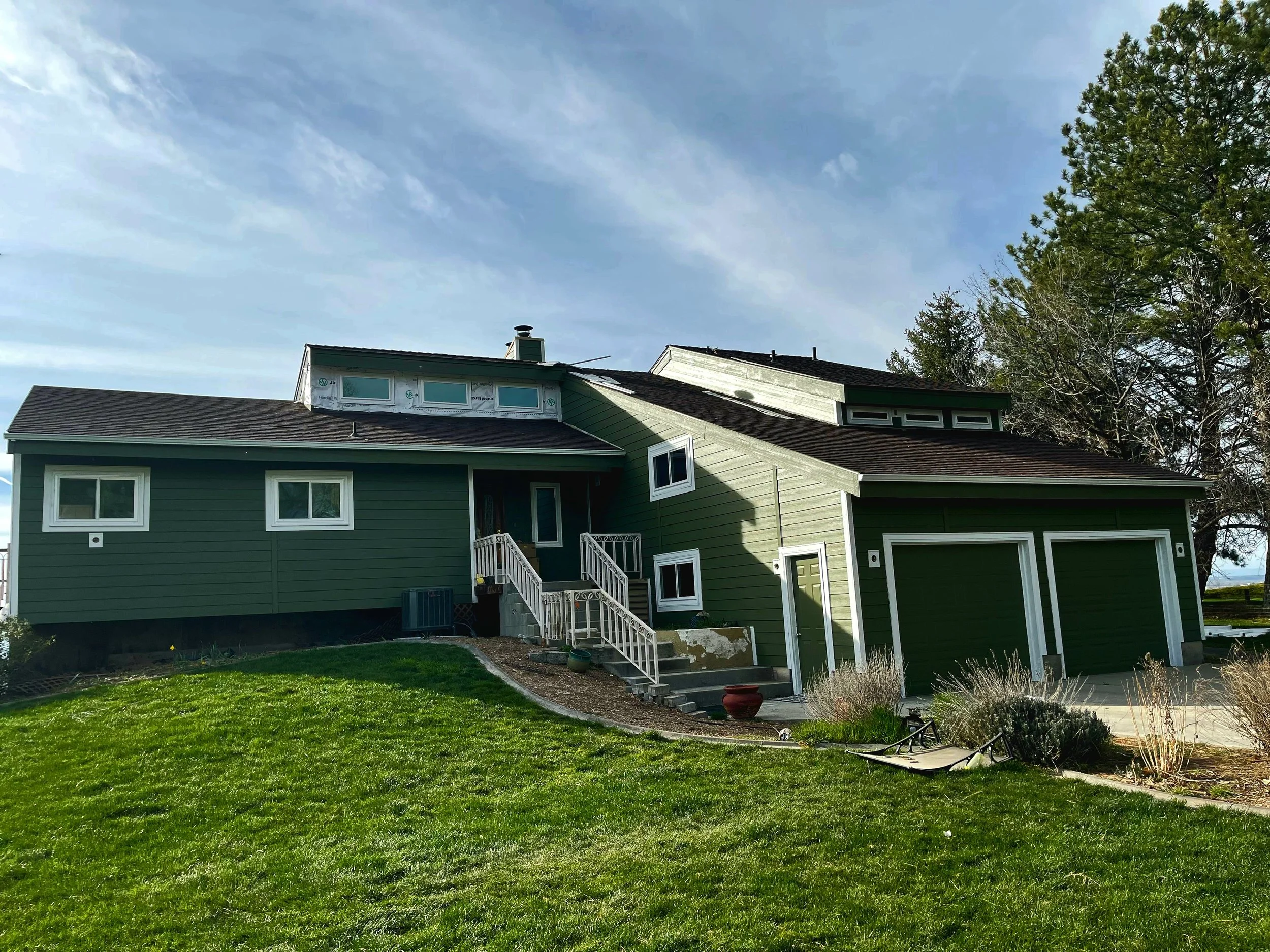 A partly under construction green house with a sloped roof and multiple windows, surrounded by a lush lawn, trees, and a partly cloudy sky.