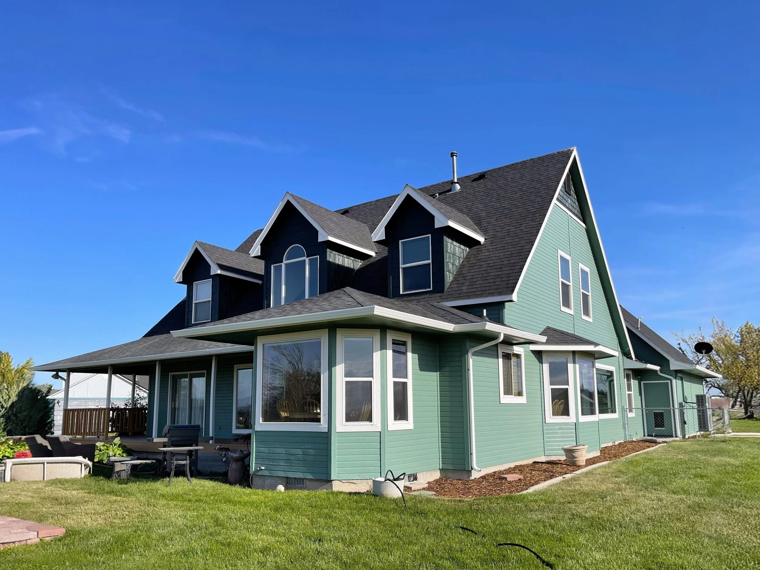 A two-story house painted teal with white trim, featuring steep gabled roofs, large windows, and a backyard with outdoor furniture and a grassy lawn under a clear blue sky.