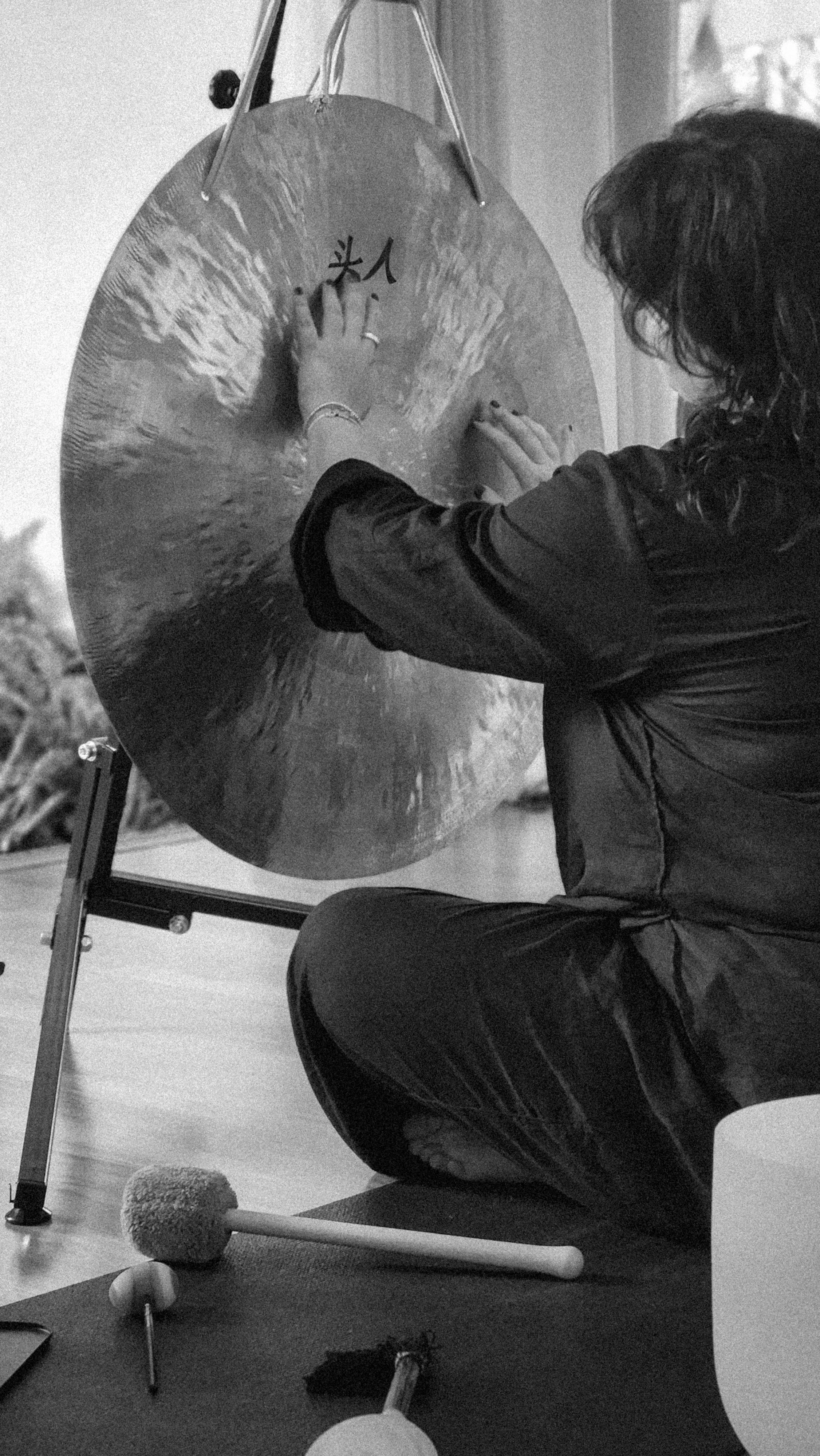 Person with curly hair playing a gong with Chinese characters on it, sitting on the floor with a mat, and various percussion instruments nearby.