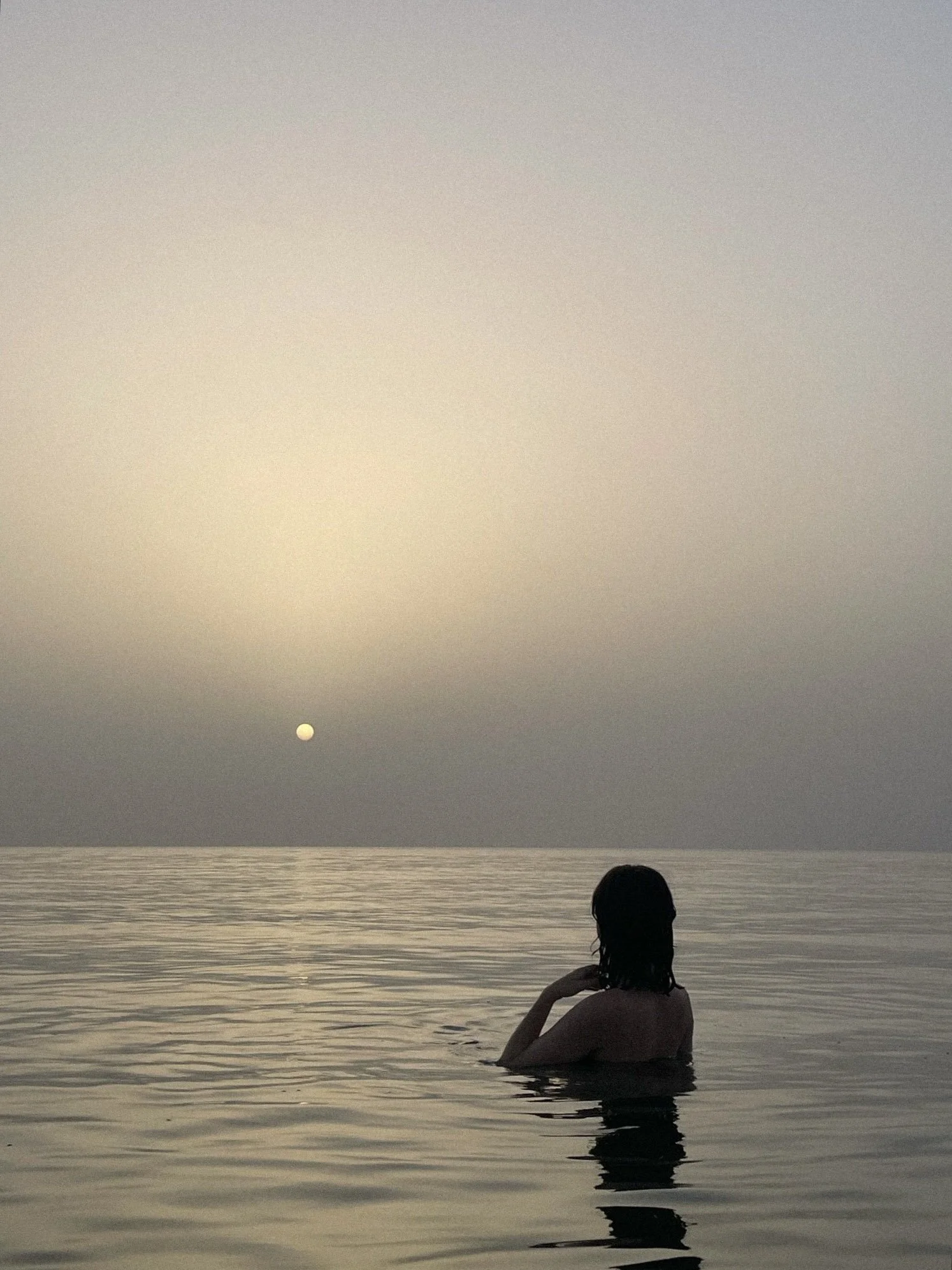 Person with long hair is sitting in calm water, facing the horizon during sunset, with a clear sky and the sun near the horizon.