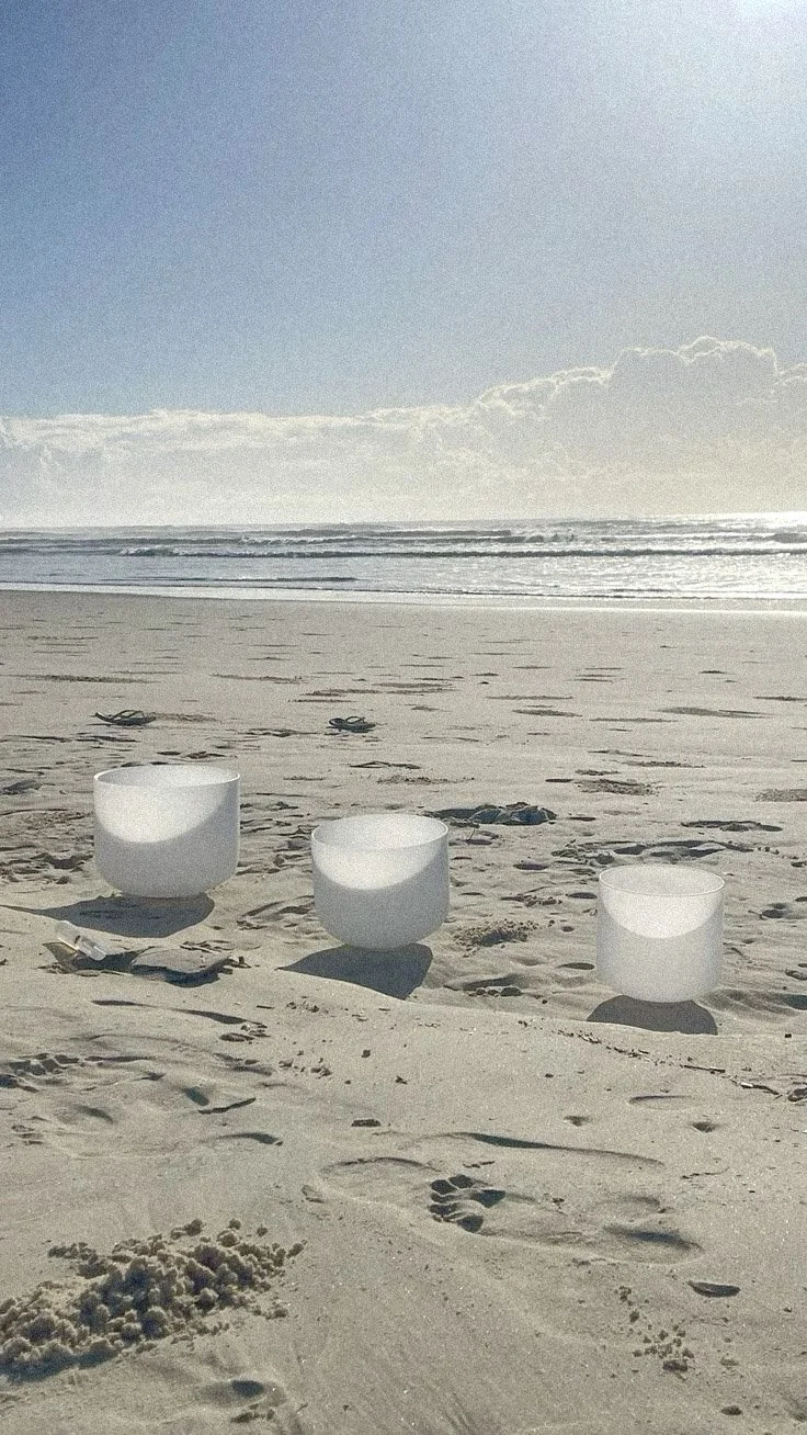 Three white crystal singing bowls of varying sizes placed on the sandy beach near the shoreline, with the ocean, waves, and a partly cloudy sky in the background.