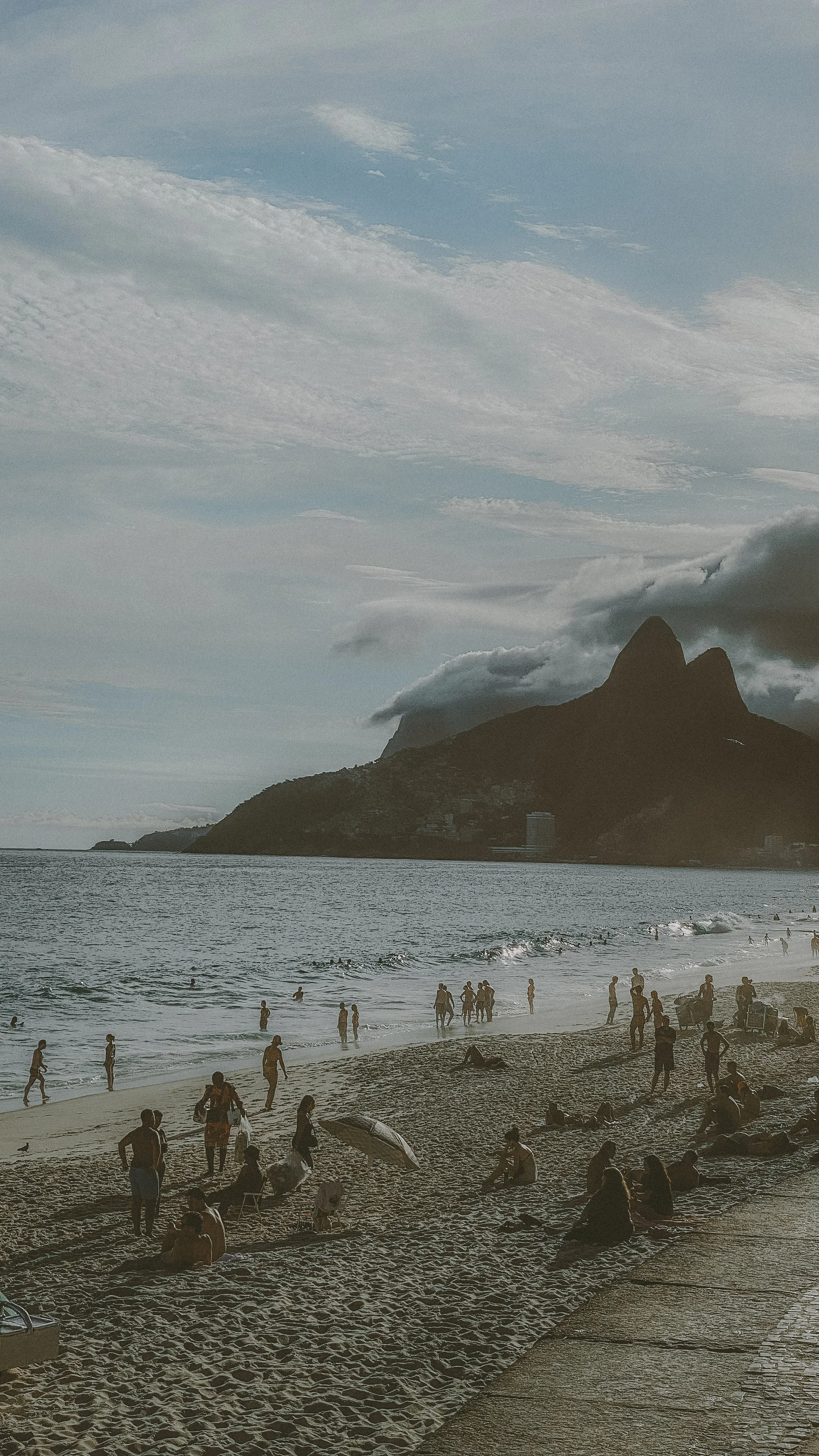 People enjoying the beach near a large mountain with clouds, with some relaxing on the sand and others in the water.