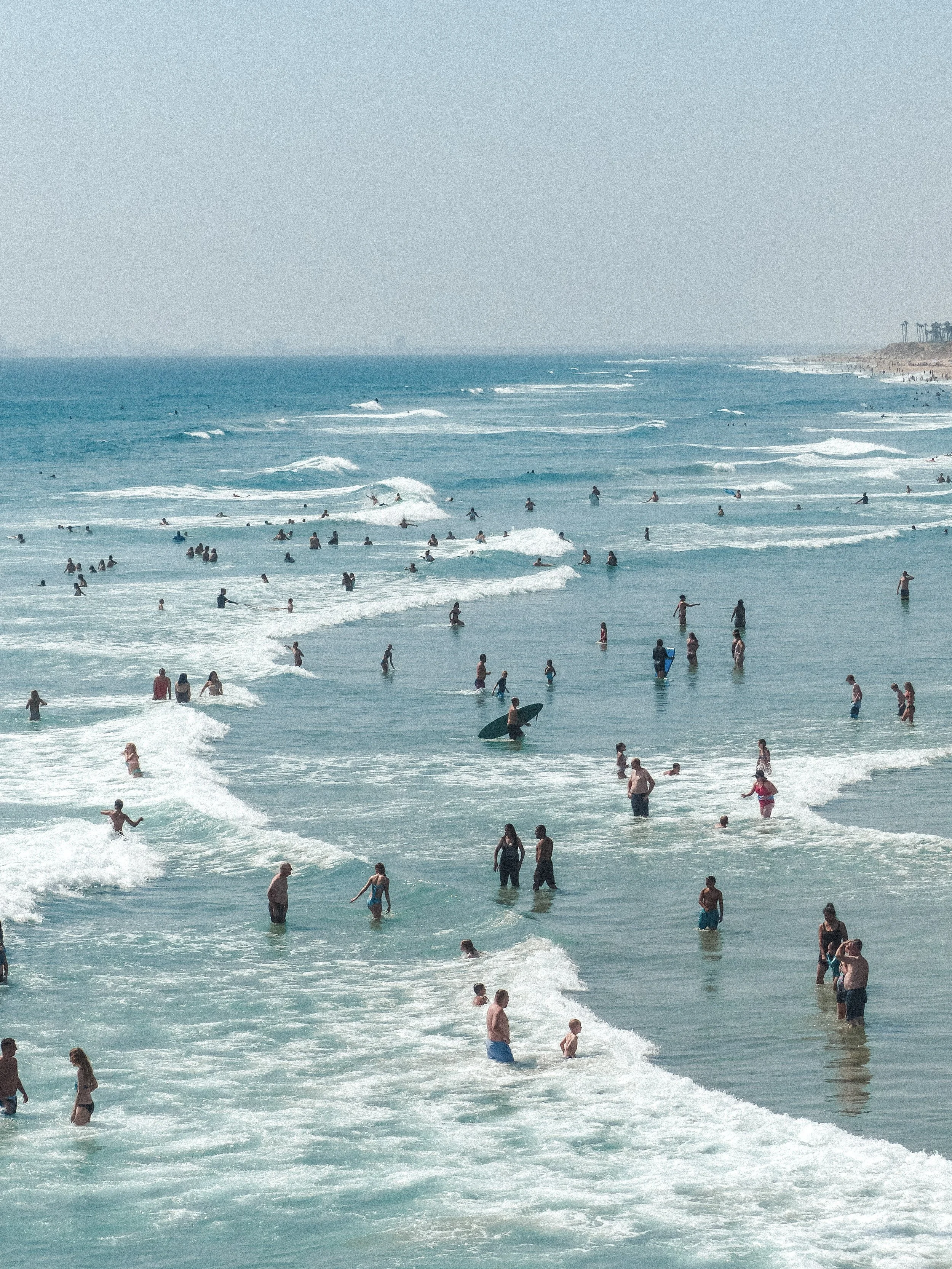 A crowded beach with many people swimming, wading, and surfing in the ocean on a sunny day.